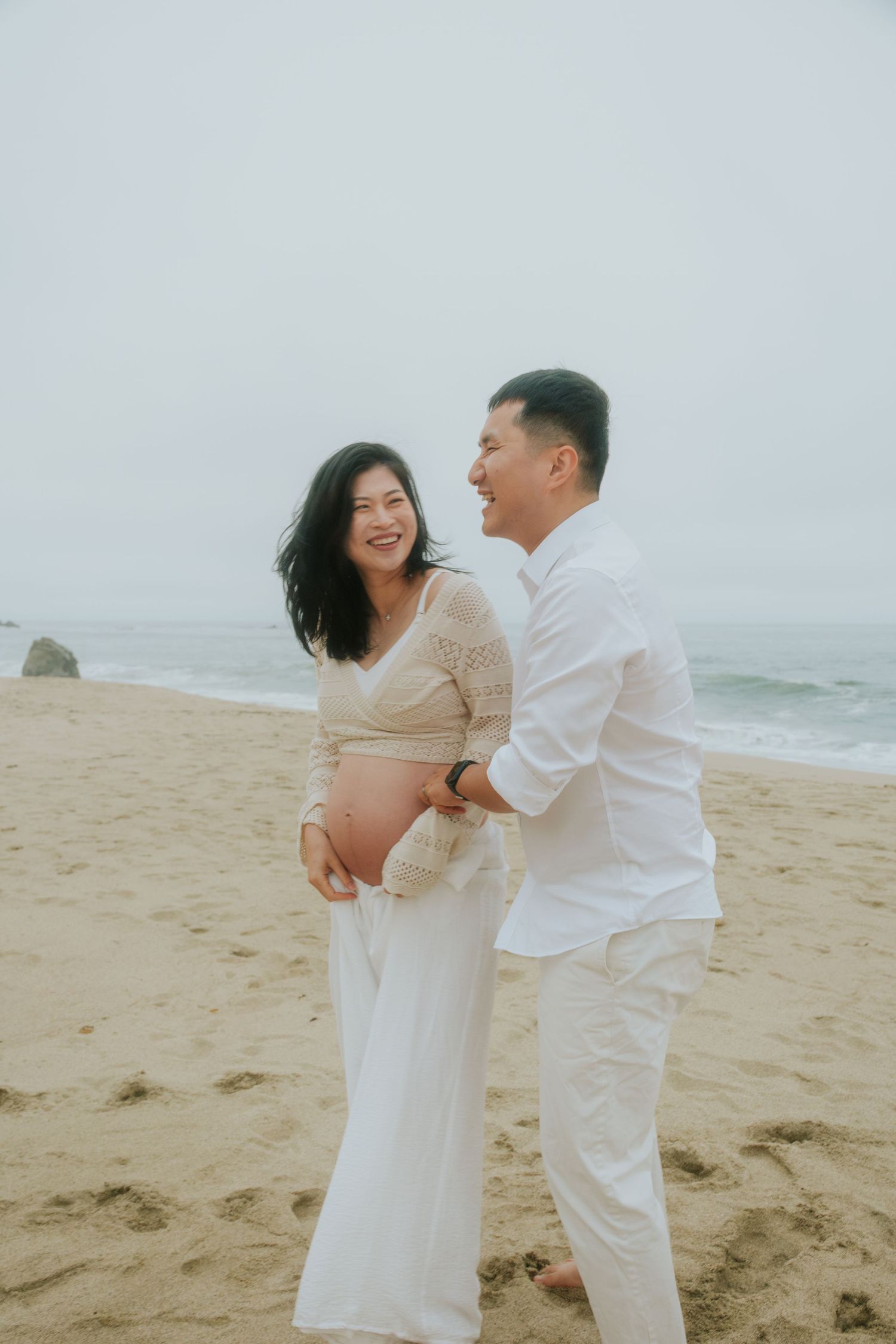 Romantic couple in white clothing embrace and laugh together during a beach photoshoot.
