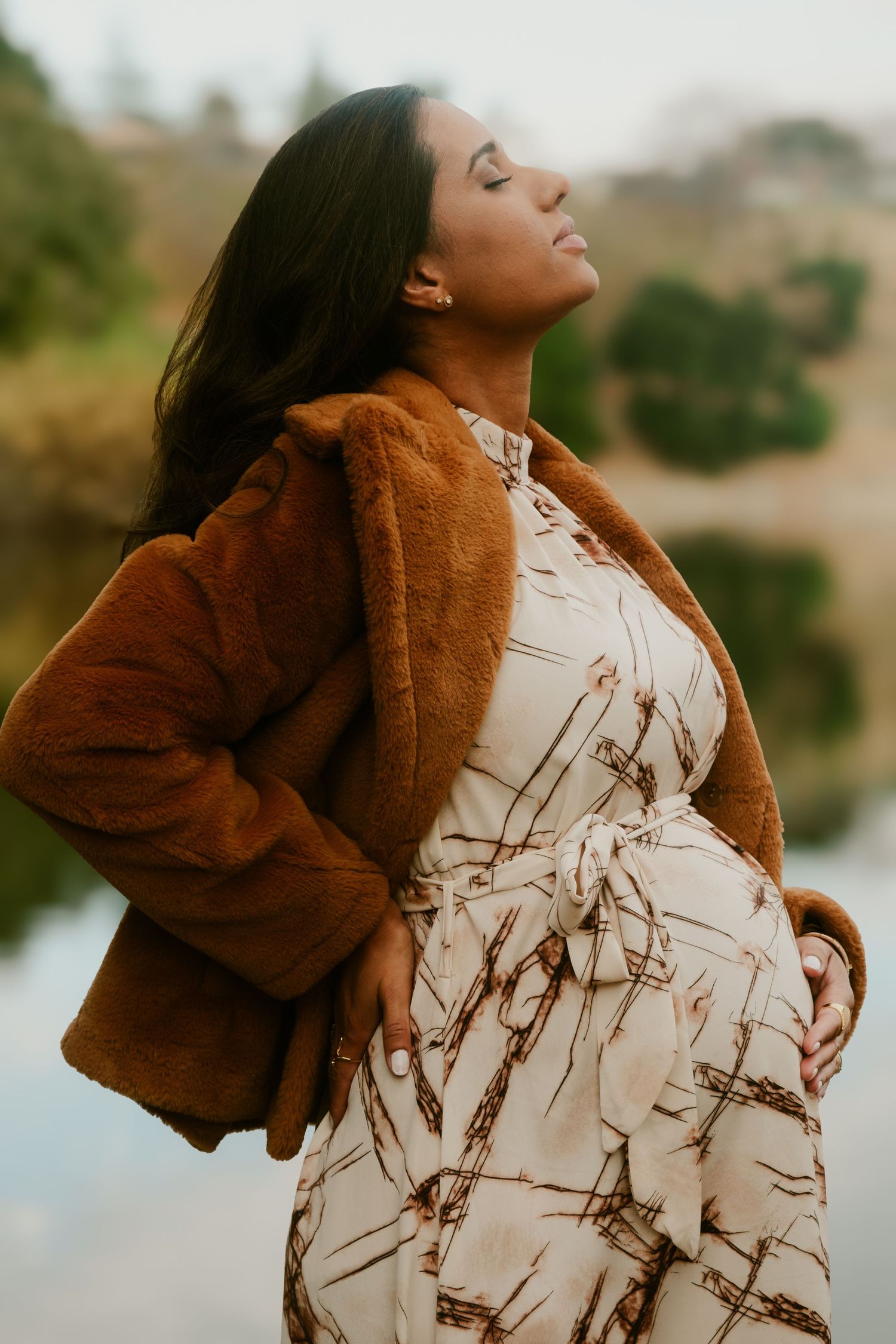 Person wearing brown coat and patterned dress poses against natural backdrop.