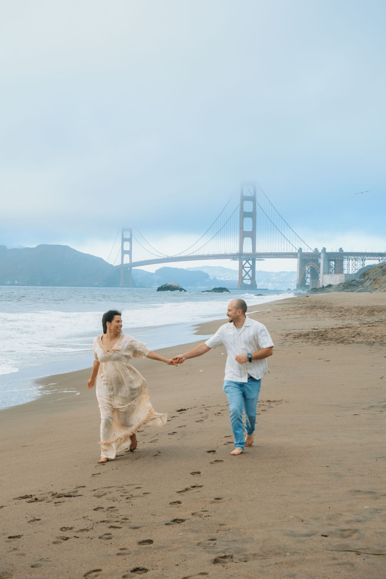 Couple holding hands while dancing on beach near Golden Gate Bridge.