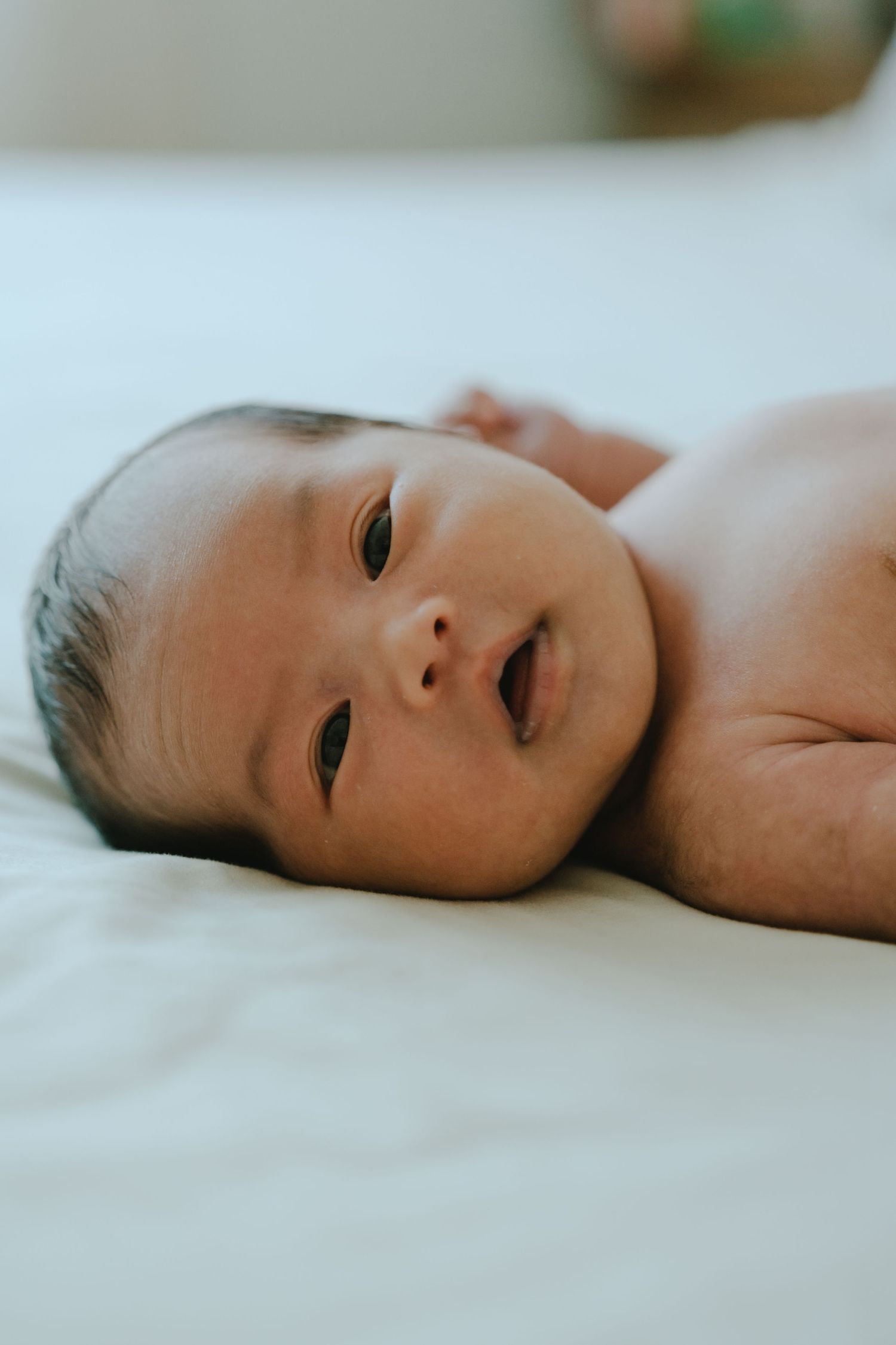 A newborn baby lying on a white surface, looking up with dark eyes and dark hair, showing chubby cheeks.