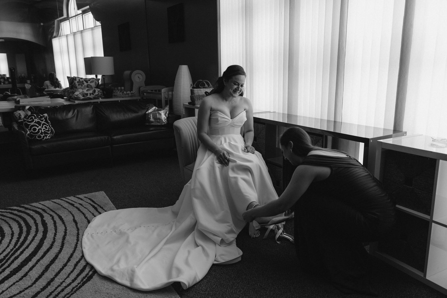 Black and white photo of a bride in a white gown being assisted by a bridesmaid in an elegant hotel suite.