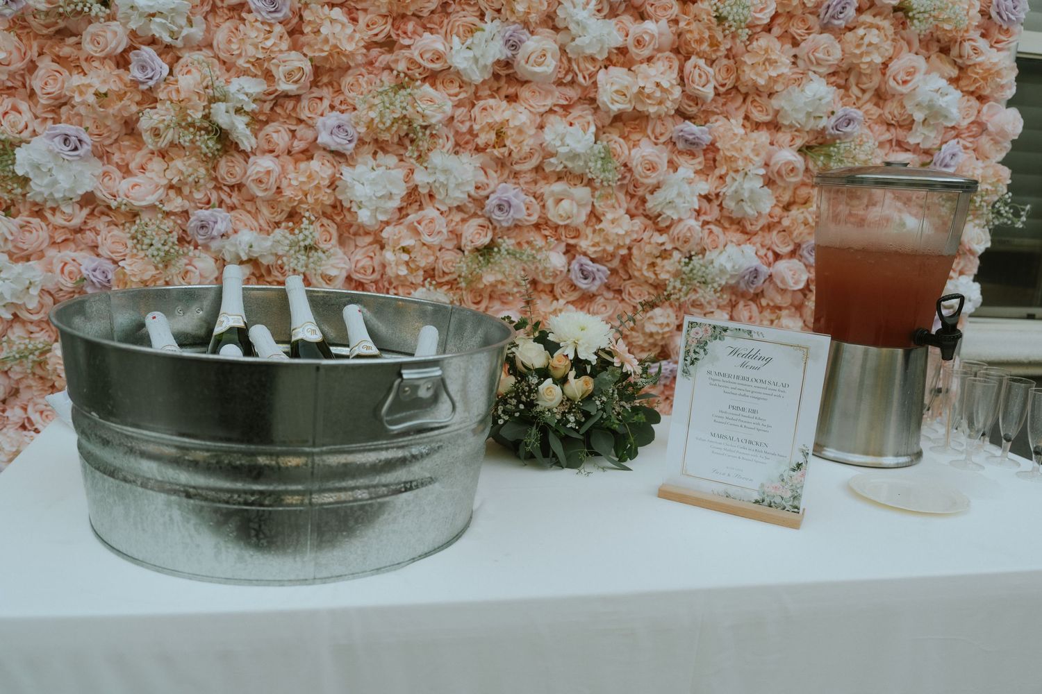 Elegant wedding drink station with champagne bucket, floral wall backdrop, and a beverage dispenser on a white table.