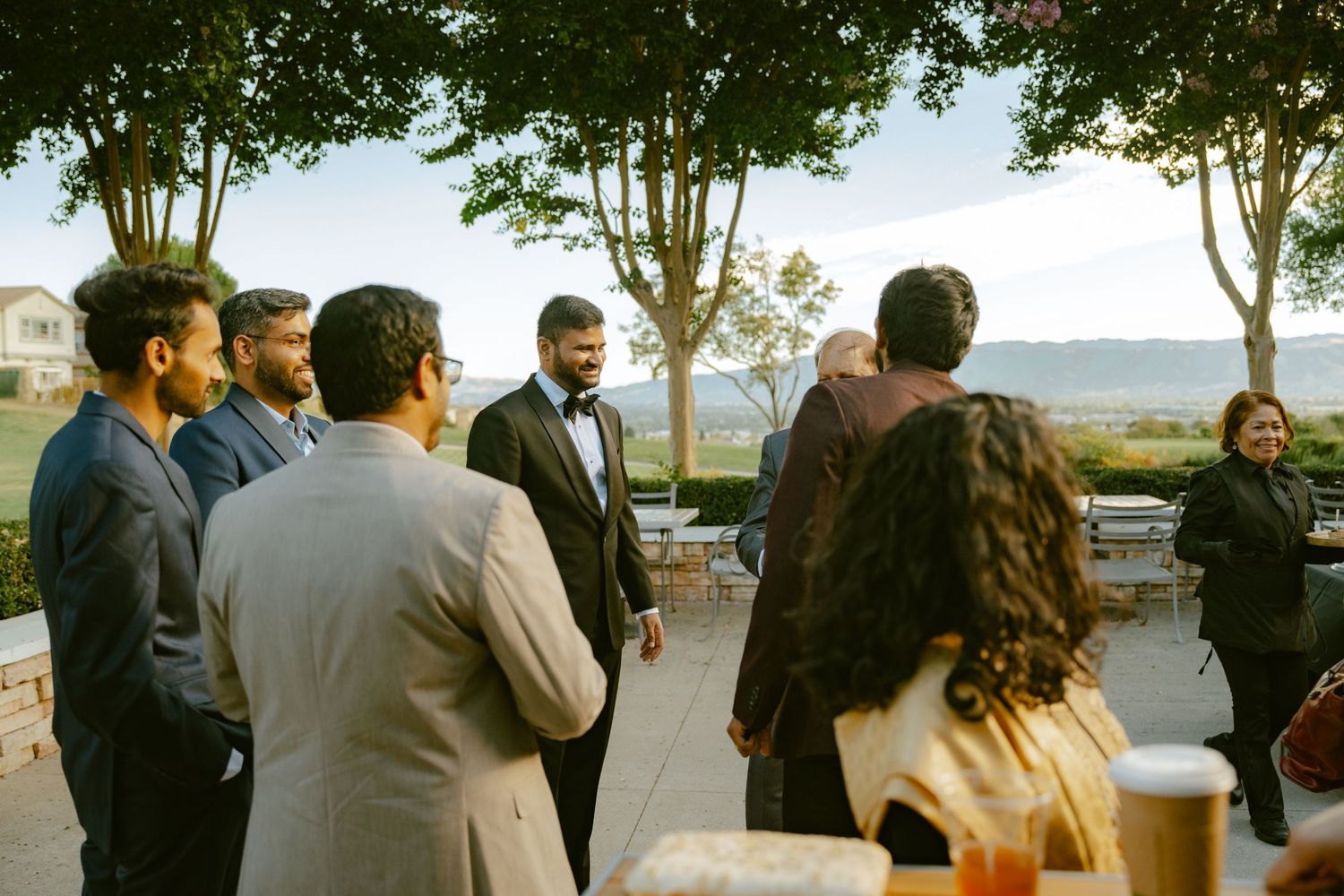 Groom in tuxedo greeting guests outdoors at elegant wedding reception with scenic mountain views.