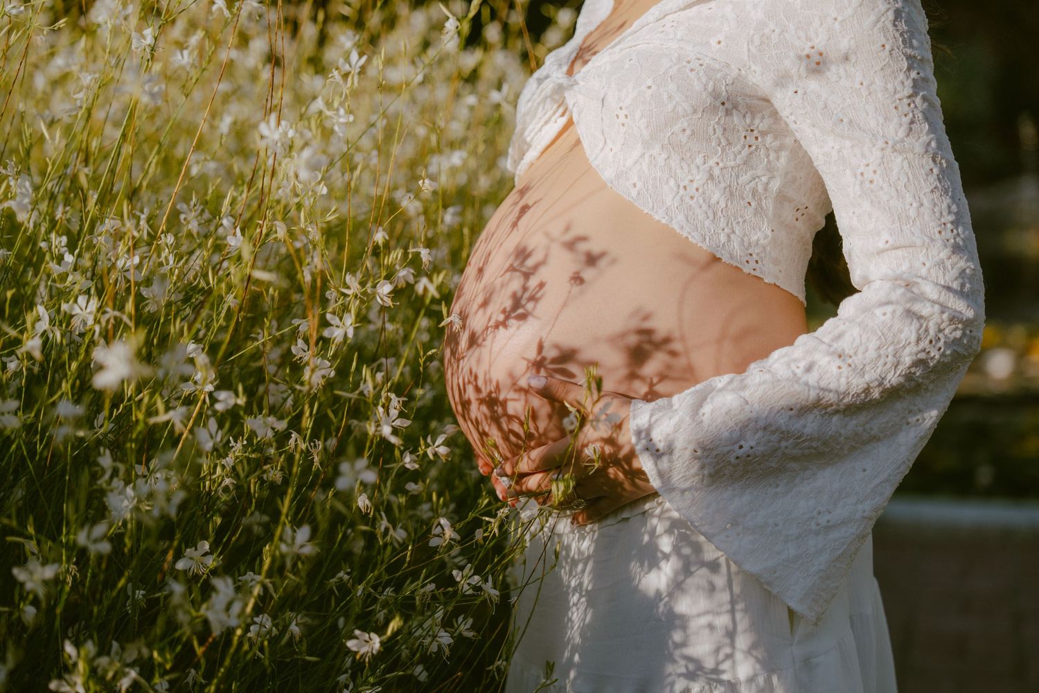Pregnant woman in white lace crop top standing in sunlit daisy field, belly exposed with flower shadows.