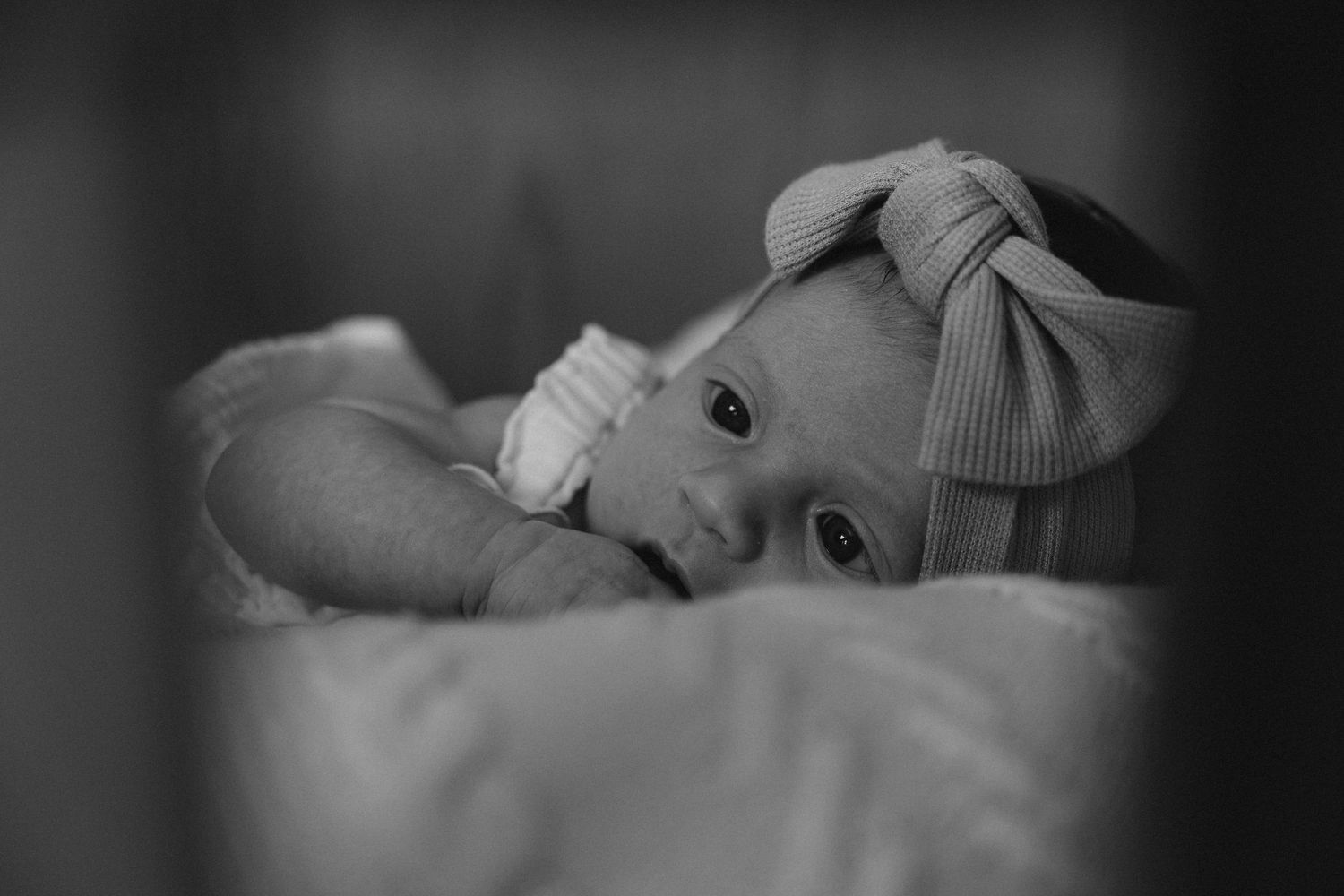 Black and white photo of a newborn baby wearing a bow headband, lying peacefully on a soft blanket.