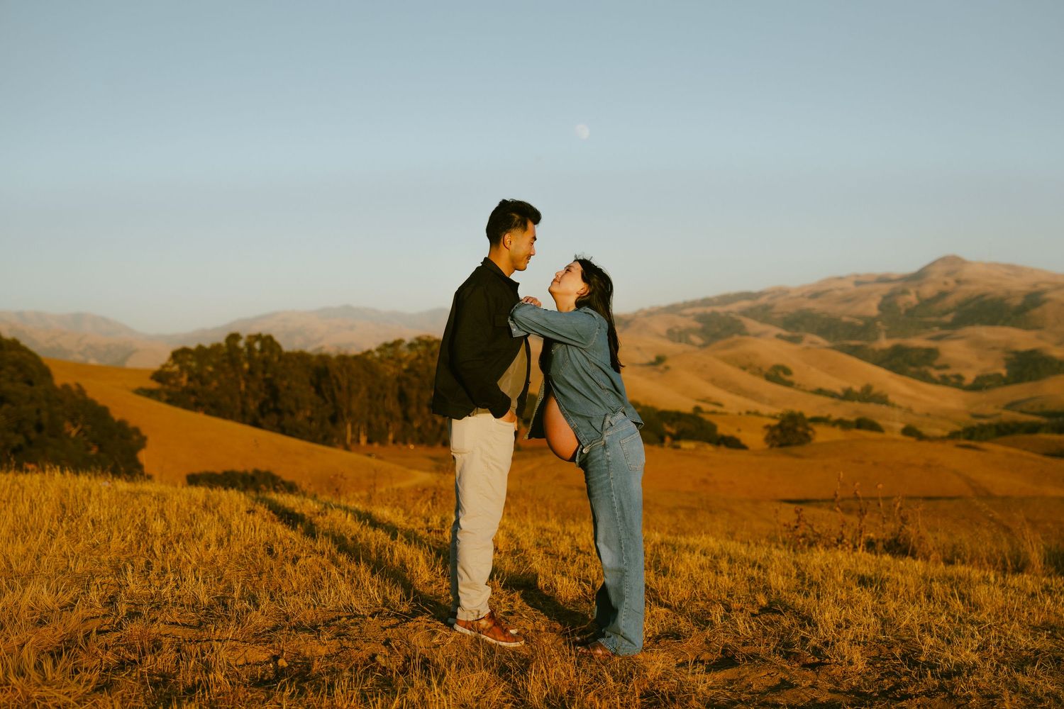 A couple shares a romantic moment in golden hills at sunset, with rolling mountains in the background.