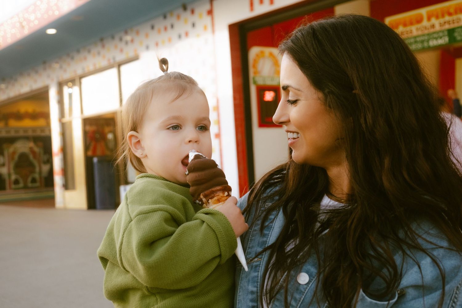 A young mother holds her toddler eating a chocolate ice cream cone outside a shop.