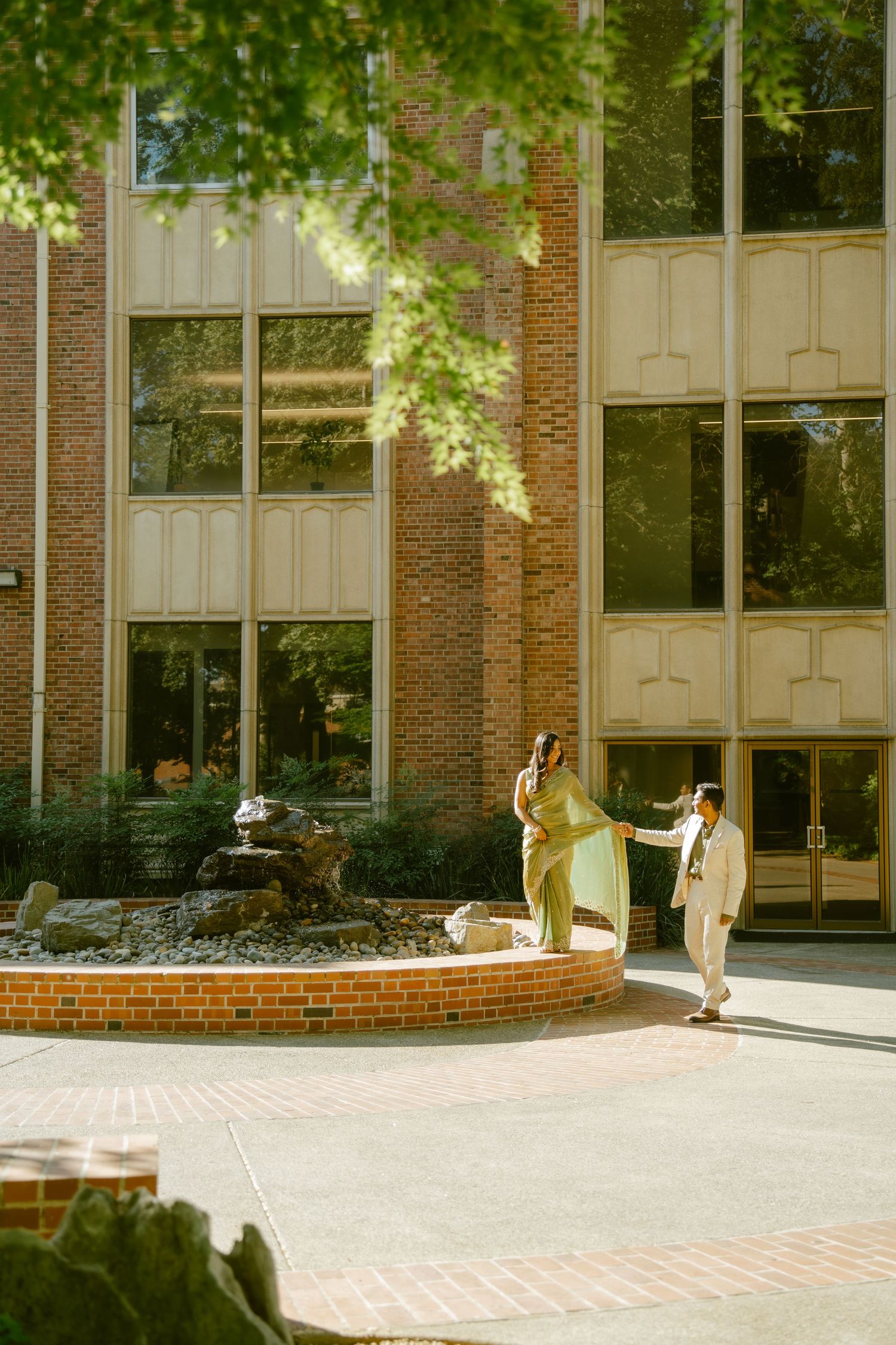 Couple in elegant attire walking near a brick fountain courtyard outside a classic brick building on a sunny day.
