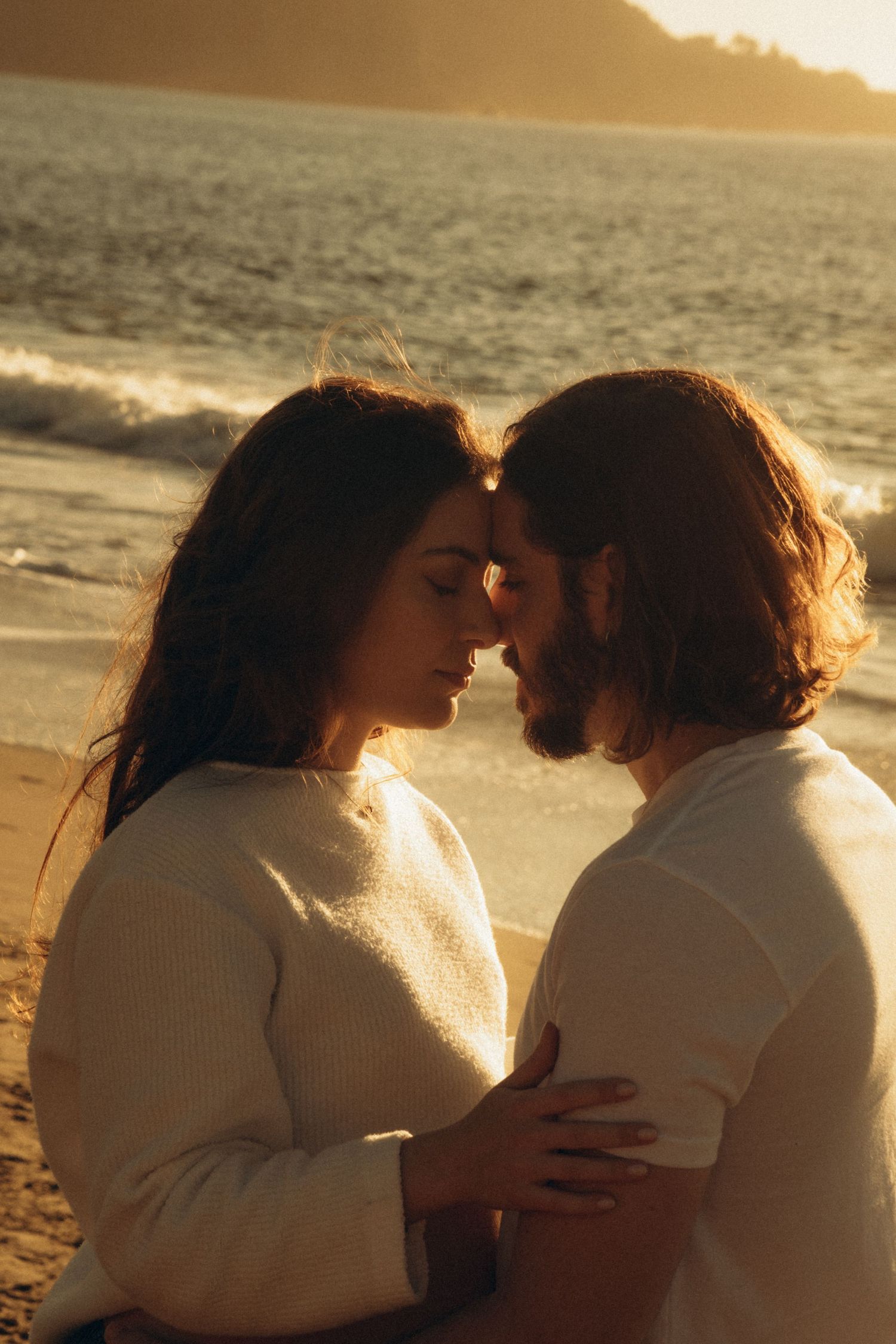 Romantic couple embracing on a beach at golden sunset, foreheads touching, wearing white sweaters by ocean waves.