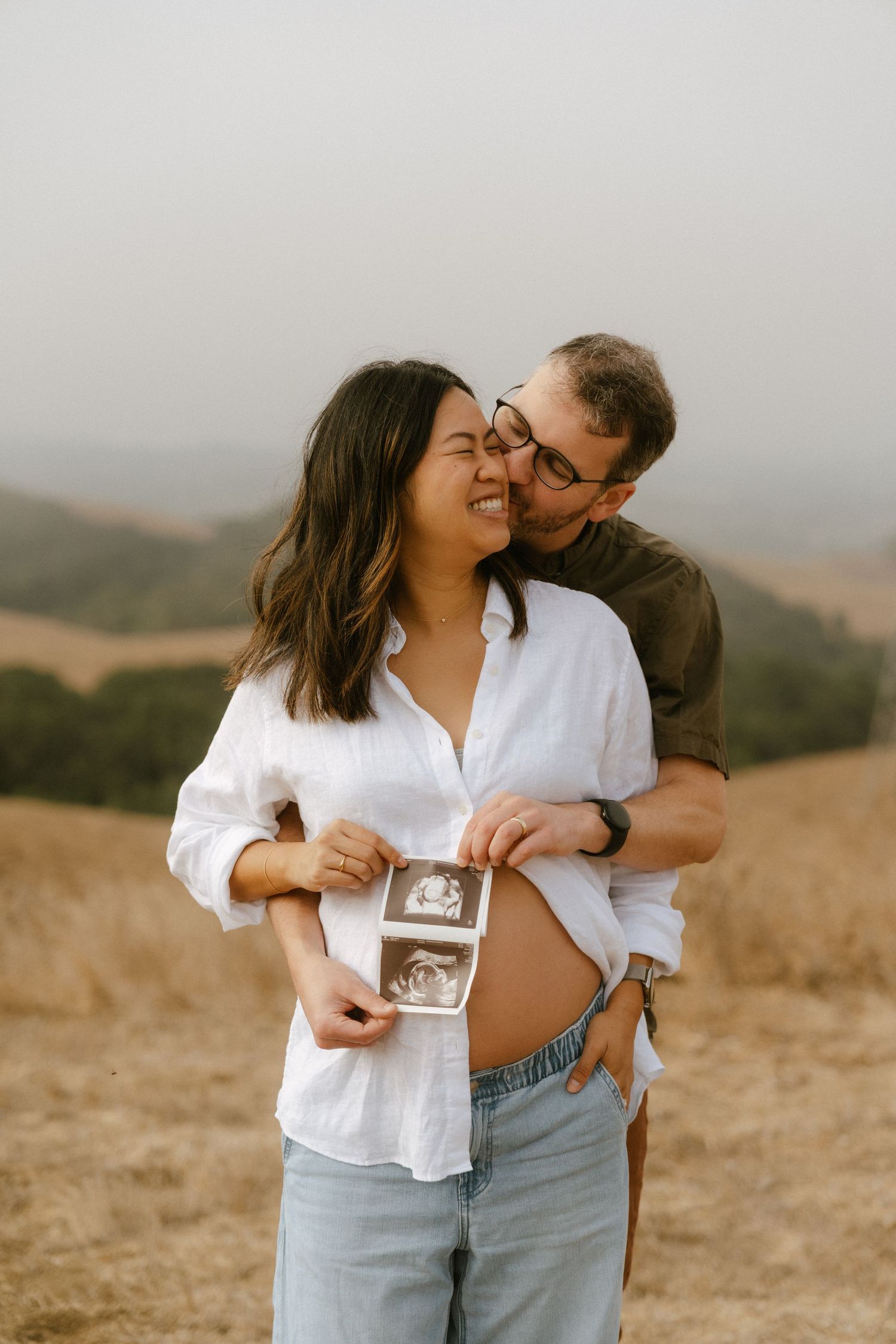 Happy couple sharing a tender moment outdoors, holding ultrasound photo while kissing, announcing pregnancy in golden field.