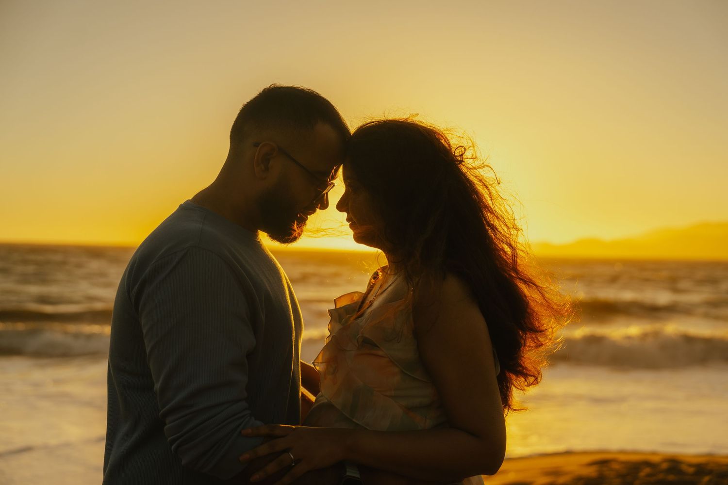 Romantic couple embracing at golden sunset on beach, silhouetted against warm orange sky and ocean waves.