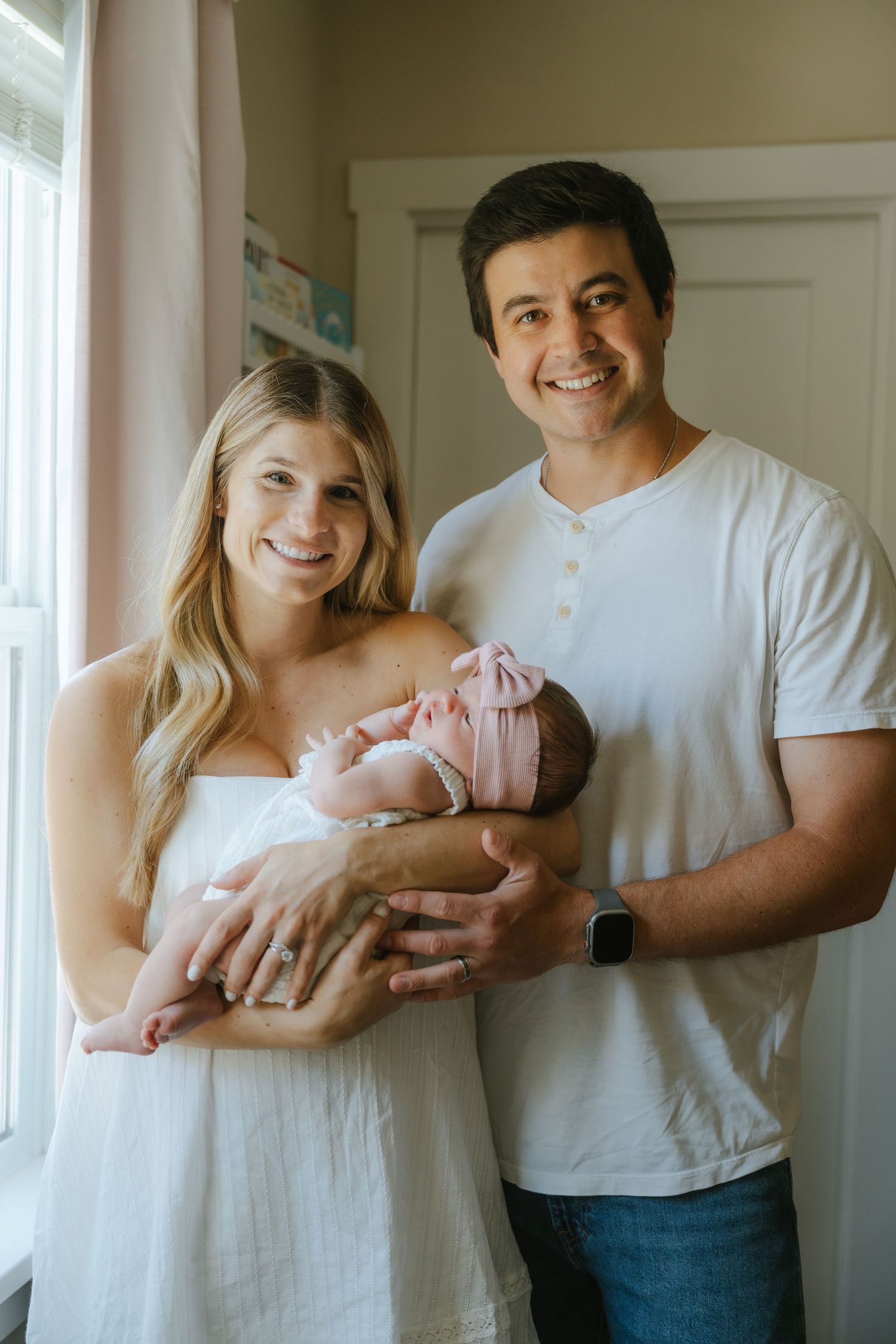 Smiling new parents holding their newborn baby in a pink headband near a bright window at home.