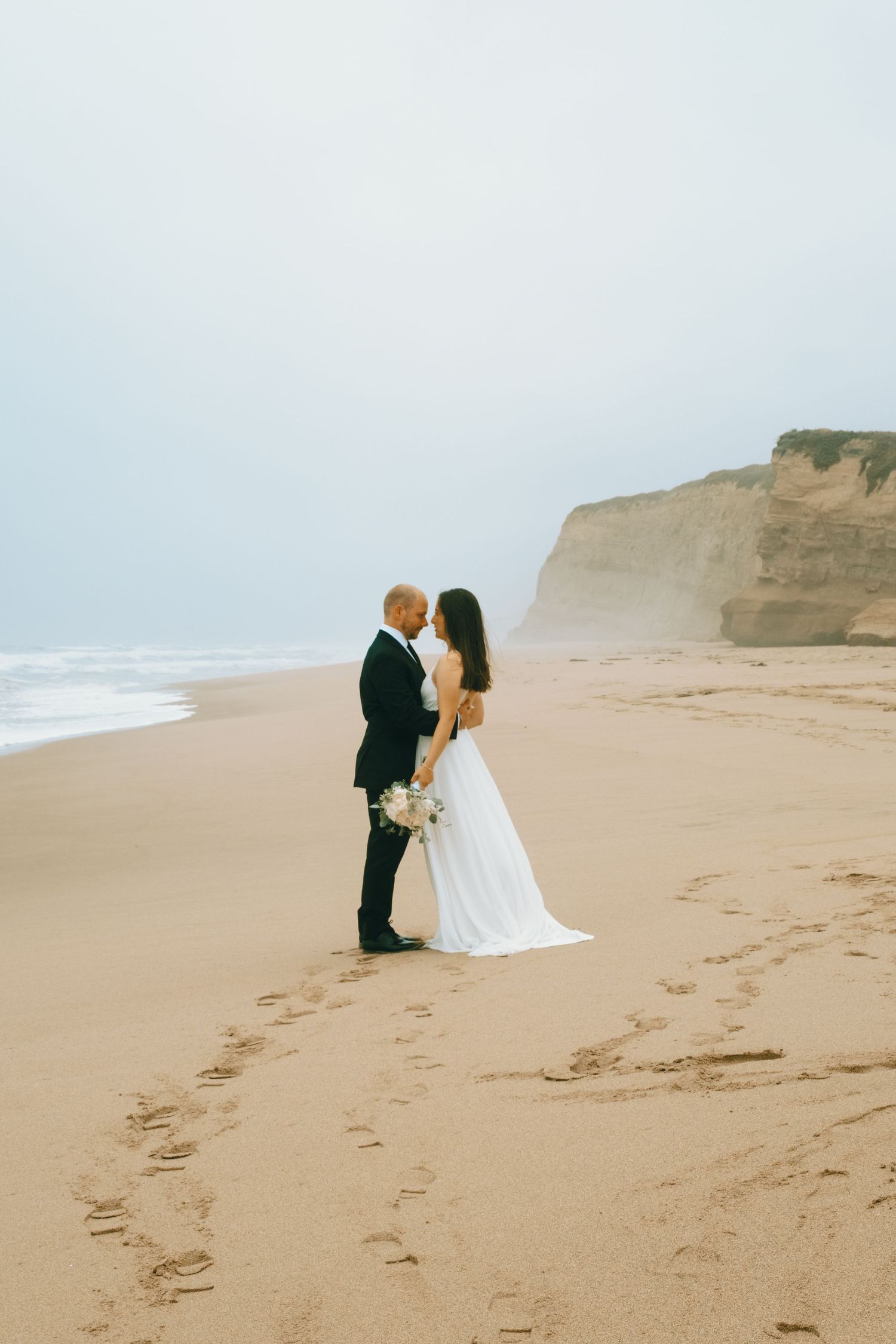 Wedding couple creates footprints in the sand during a beach photoshoot with coastal cliffs.