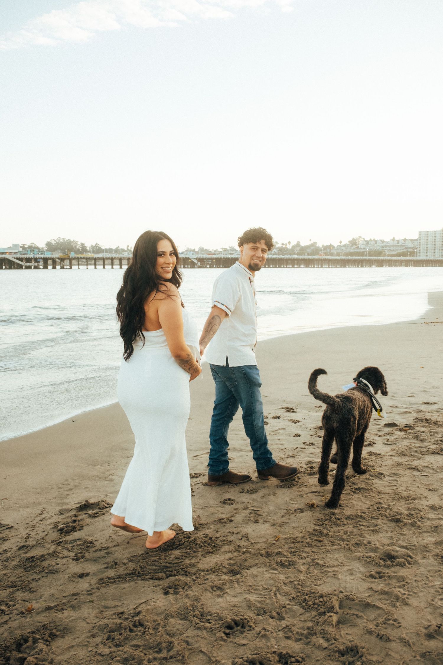 A couple walks their black dog along a scenic beach during a romantic photoshoot.
