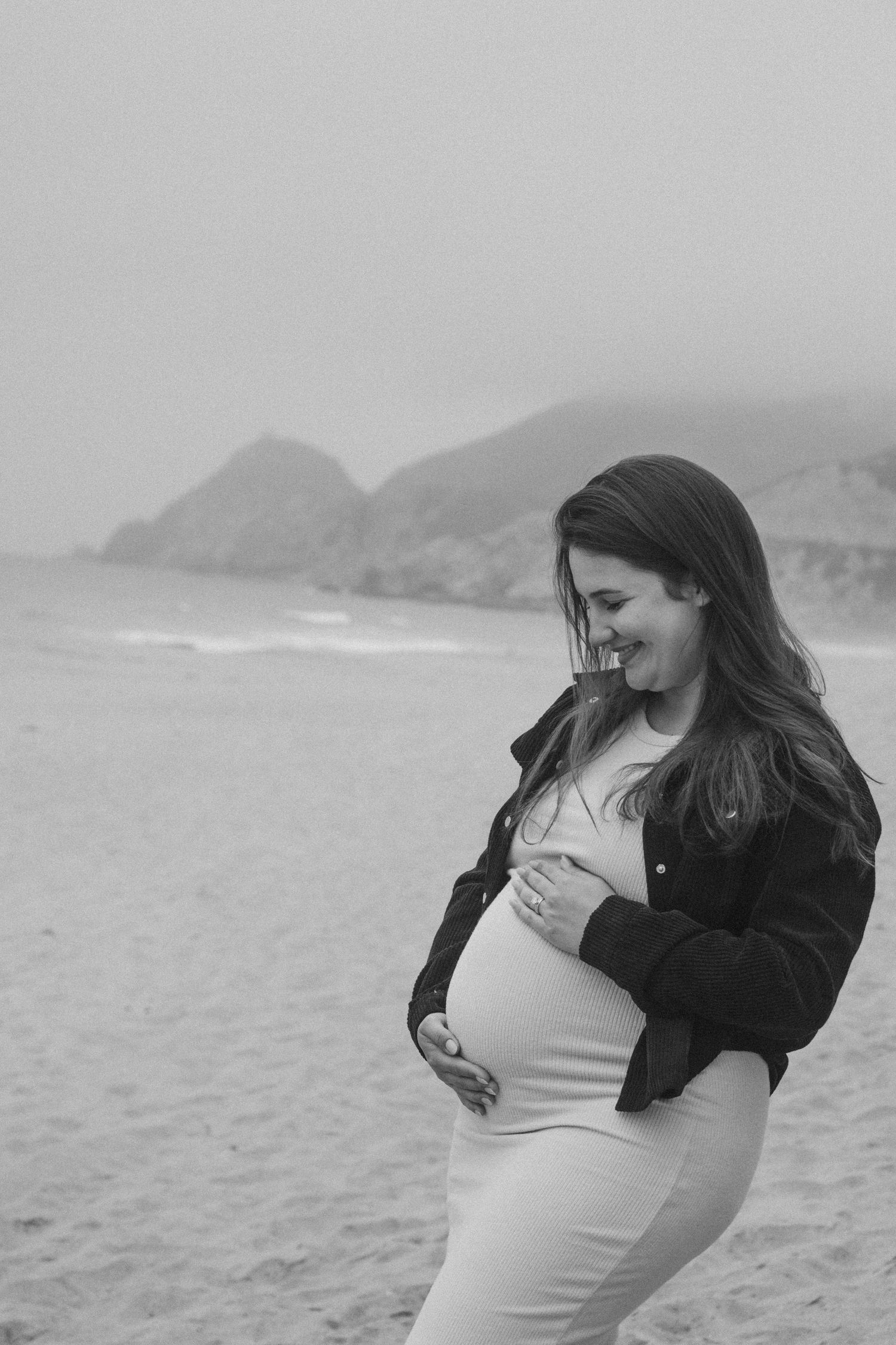 Maternity photoshoot on cloudy beach with dramatic coastal cliffs in background.