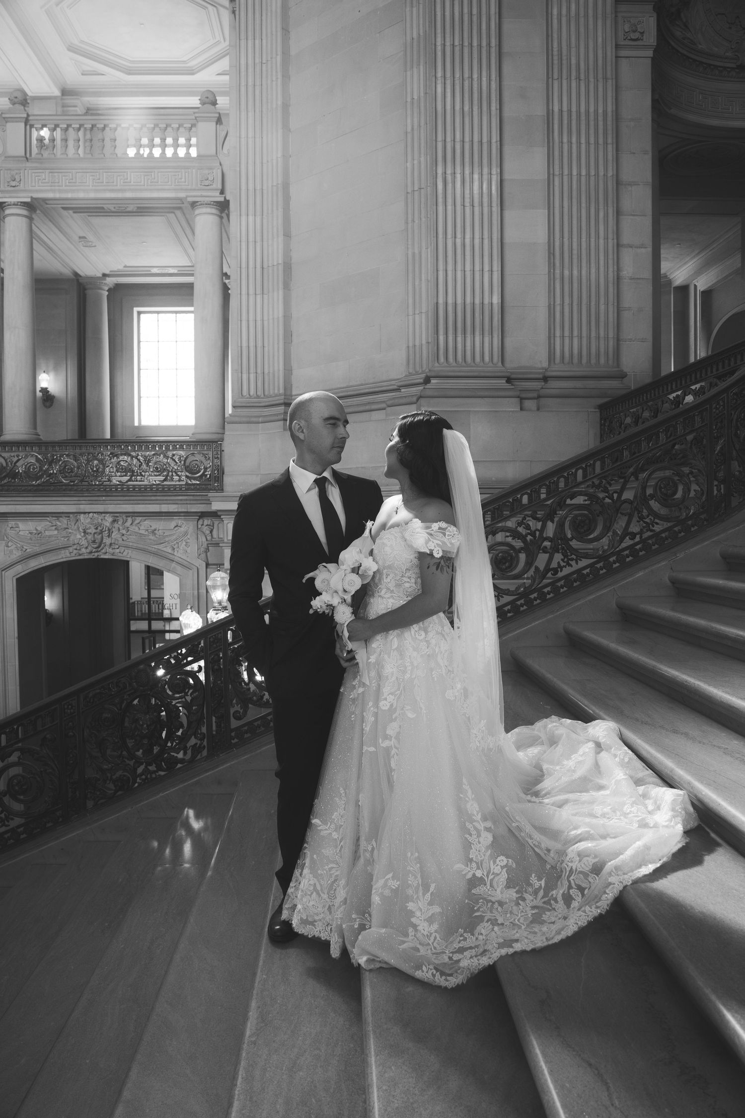 A couple poses on an elegant marble staircase with ornate iron railings.