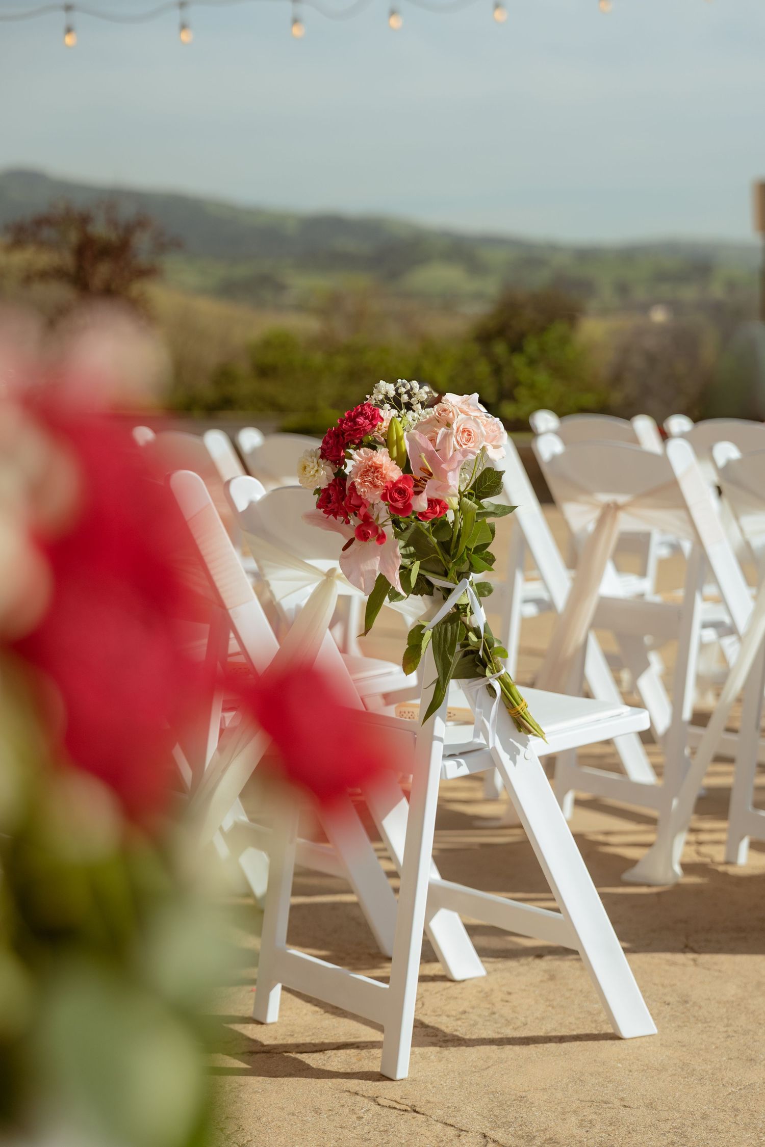 Pink and red bridal bouquet resting on white folding chairs at an outdoor wedding ceremony.