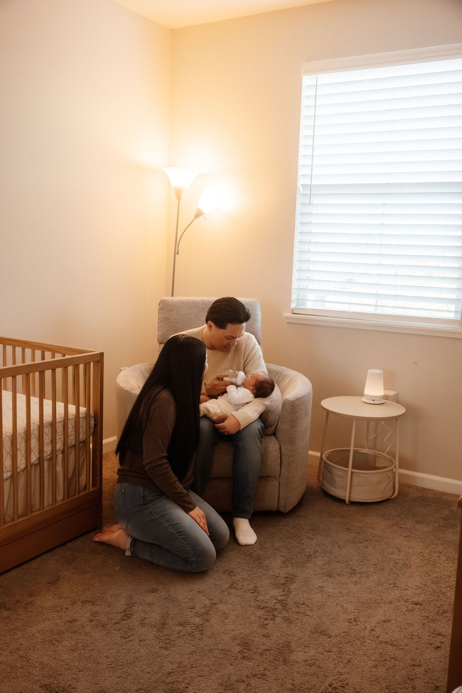 A nursery room with wooden crib, gray armchair, white side table and warm lighting from a floor lamp.