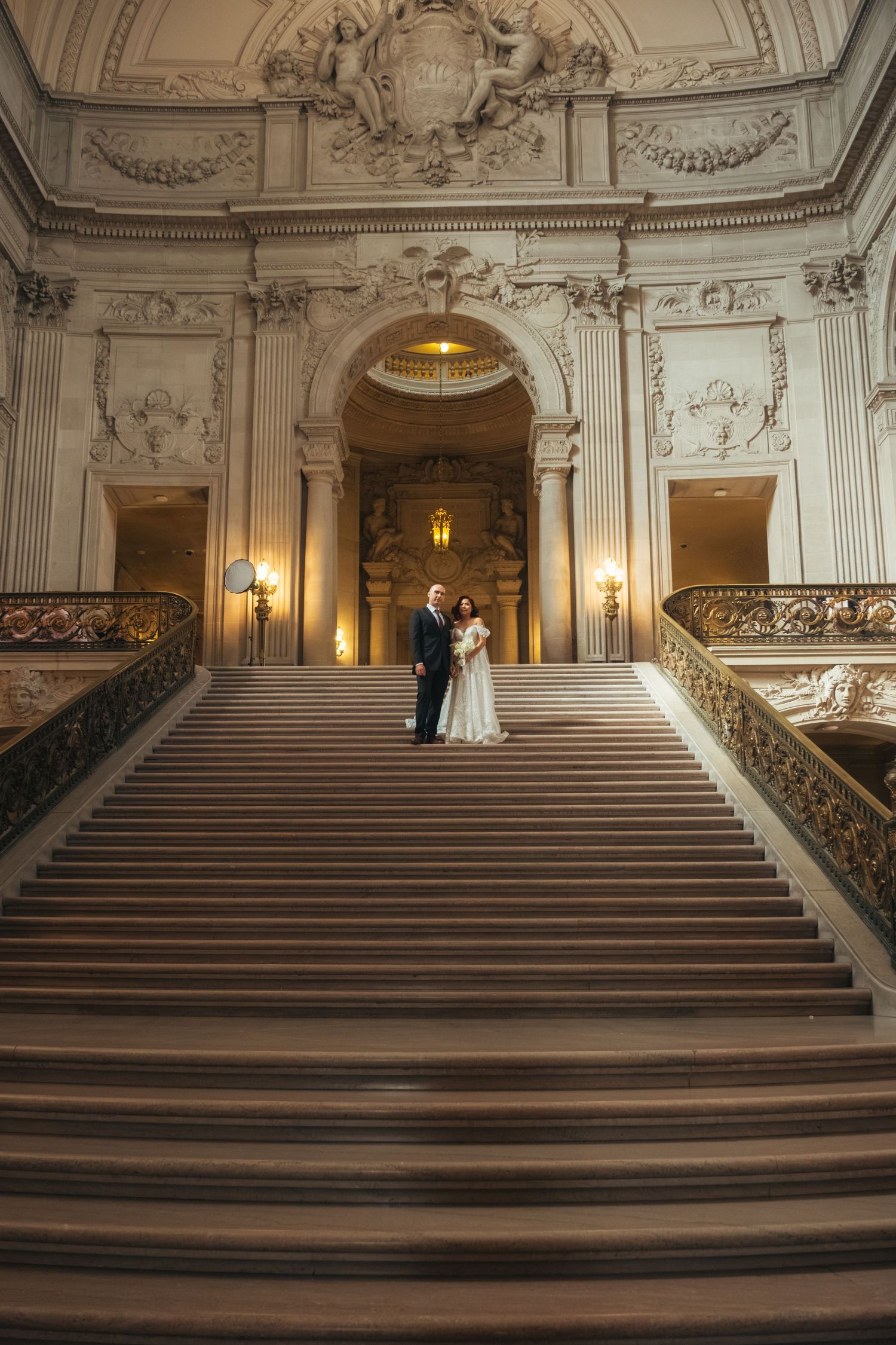 A wedding couple poses on ornate marble stairs in a luxurious classical building with dramatic lighting.