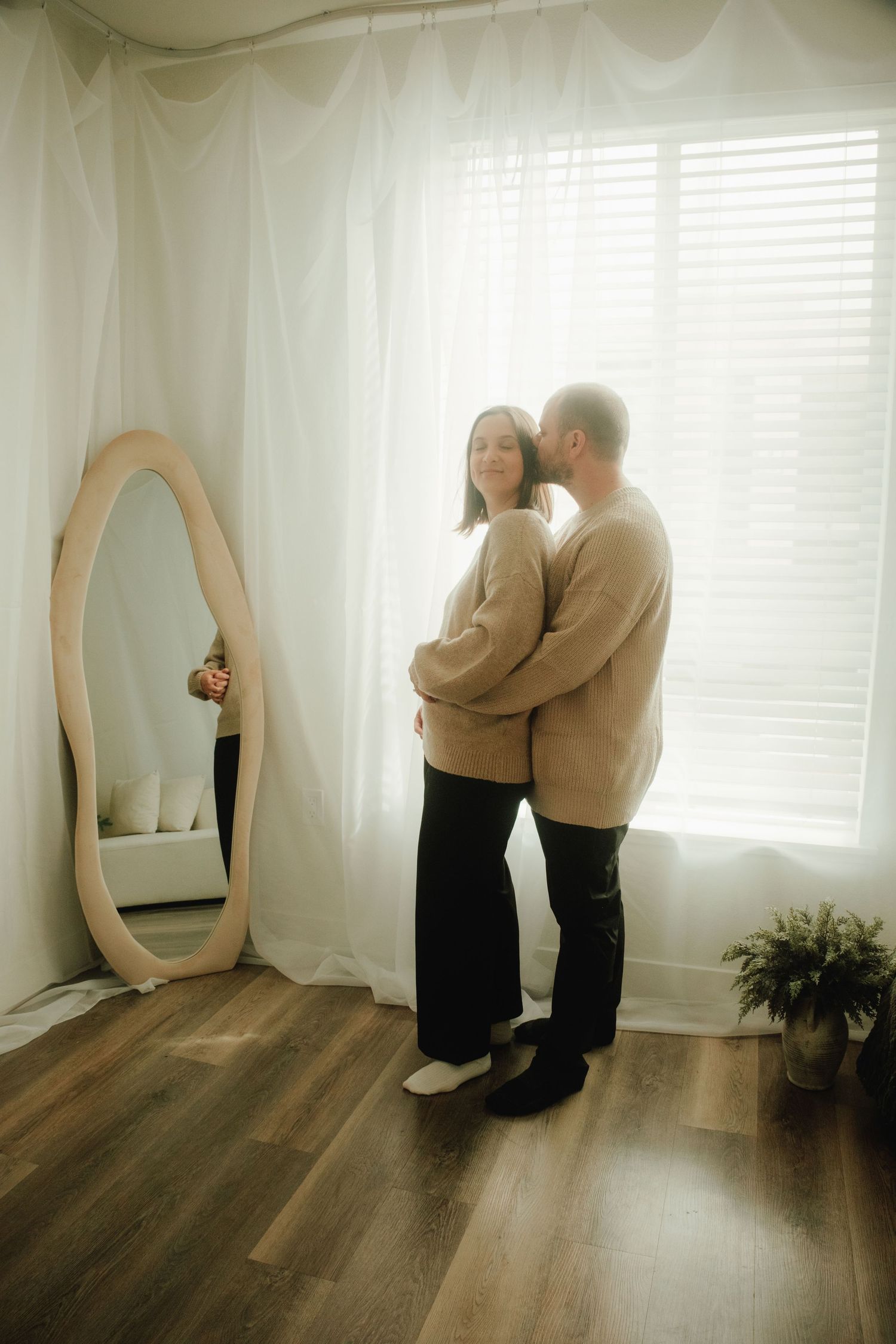 Couple in matching sweaters standing by oval mirror with Christmas decorations visible.
