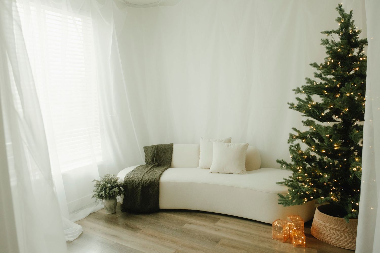A minimalist white room decorated with a lit Christmas tree and white curtains.