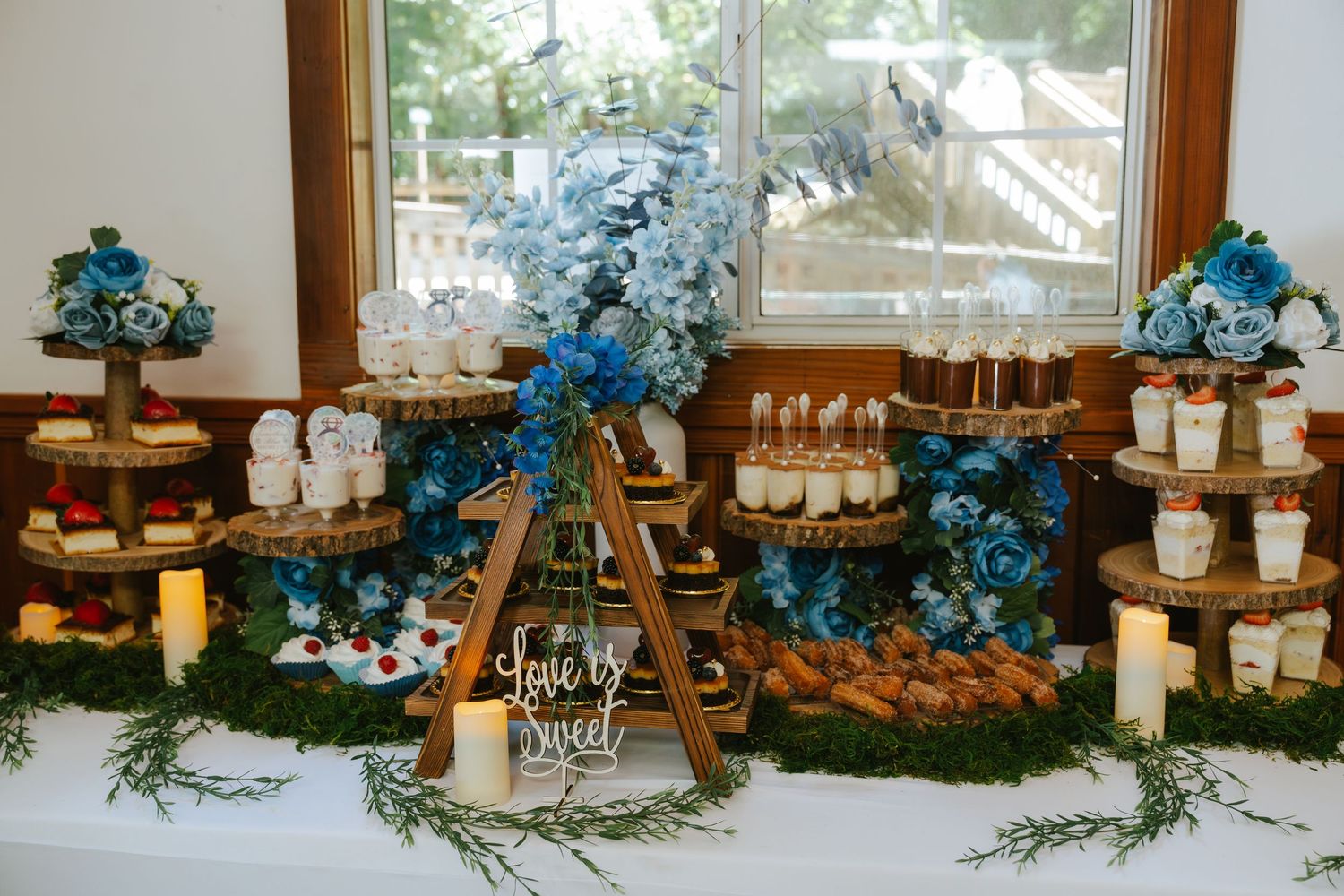 Elegant wedding dessert table with blue floral backdrop, rustic wood stands, candles, and moss accents displaying cakes and treats.