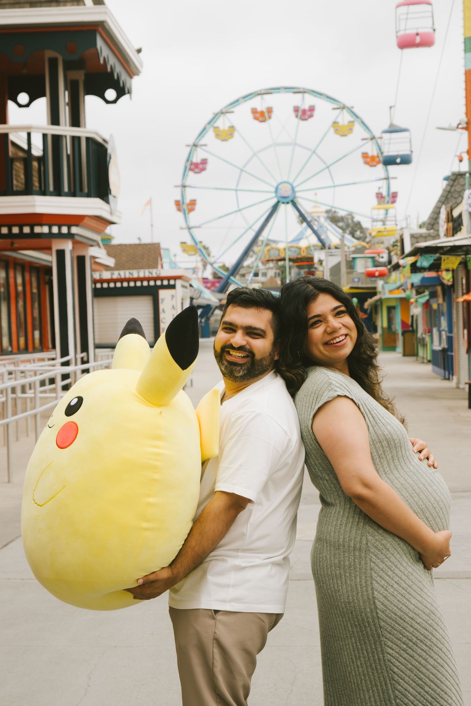 A couple poses with large yellow Pikachu plush toys at an amusement park with a Ferris wheel.