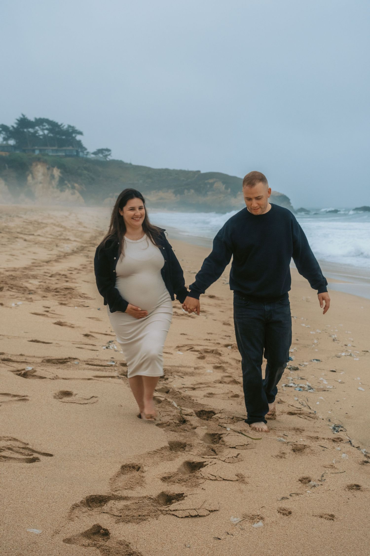 Expecting couple walking together on sandy beach leaving footprints behind.