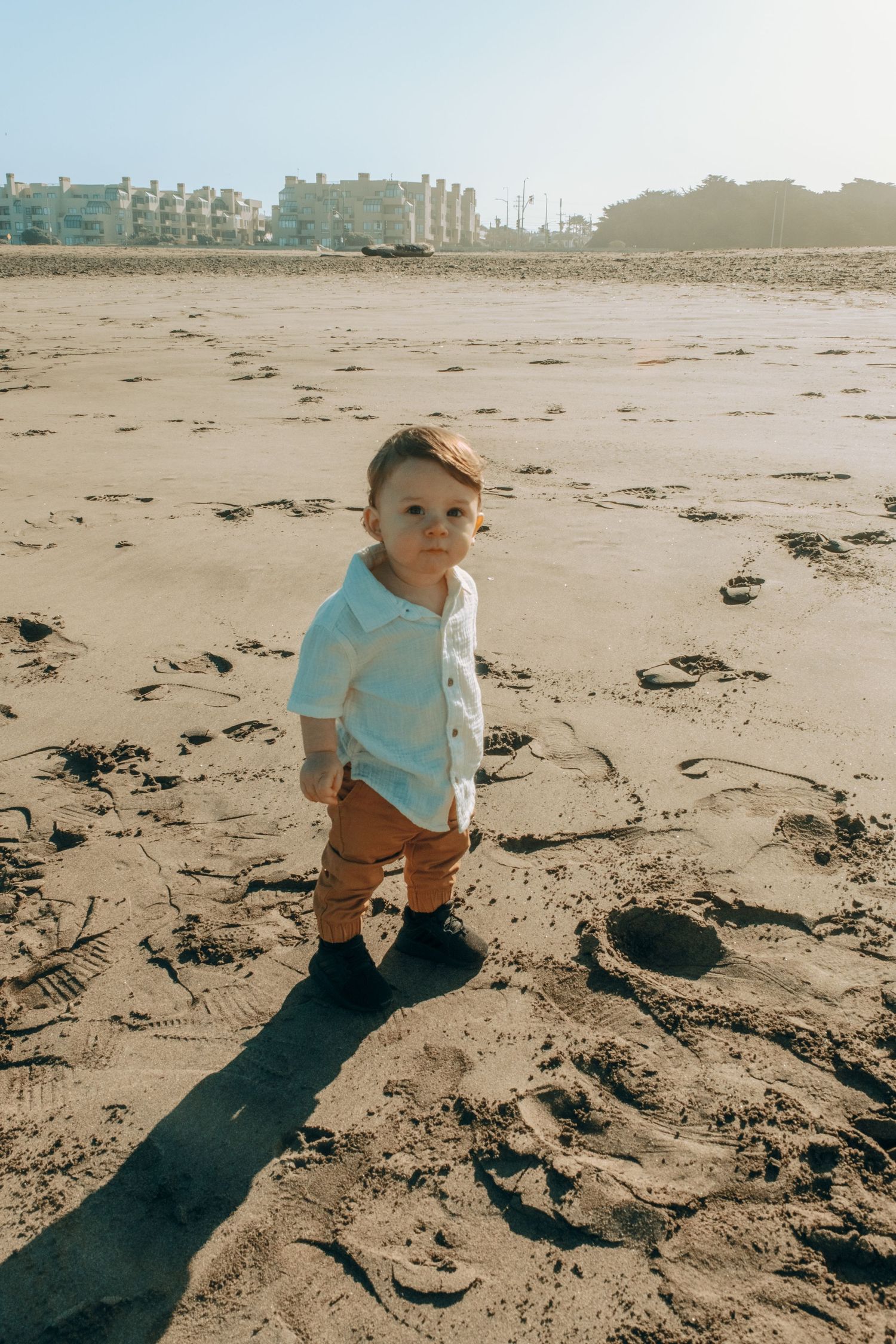 A small child explores a sandy beach wearing light blue and brown clothing on a sunny day.