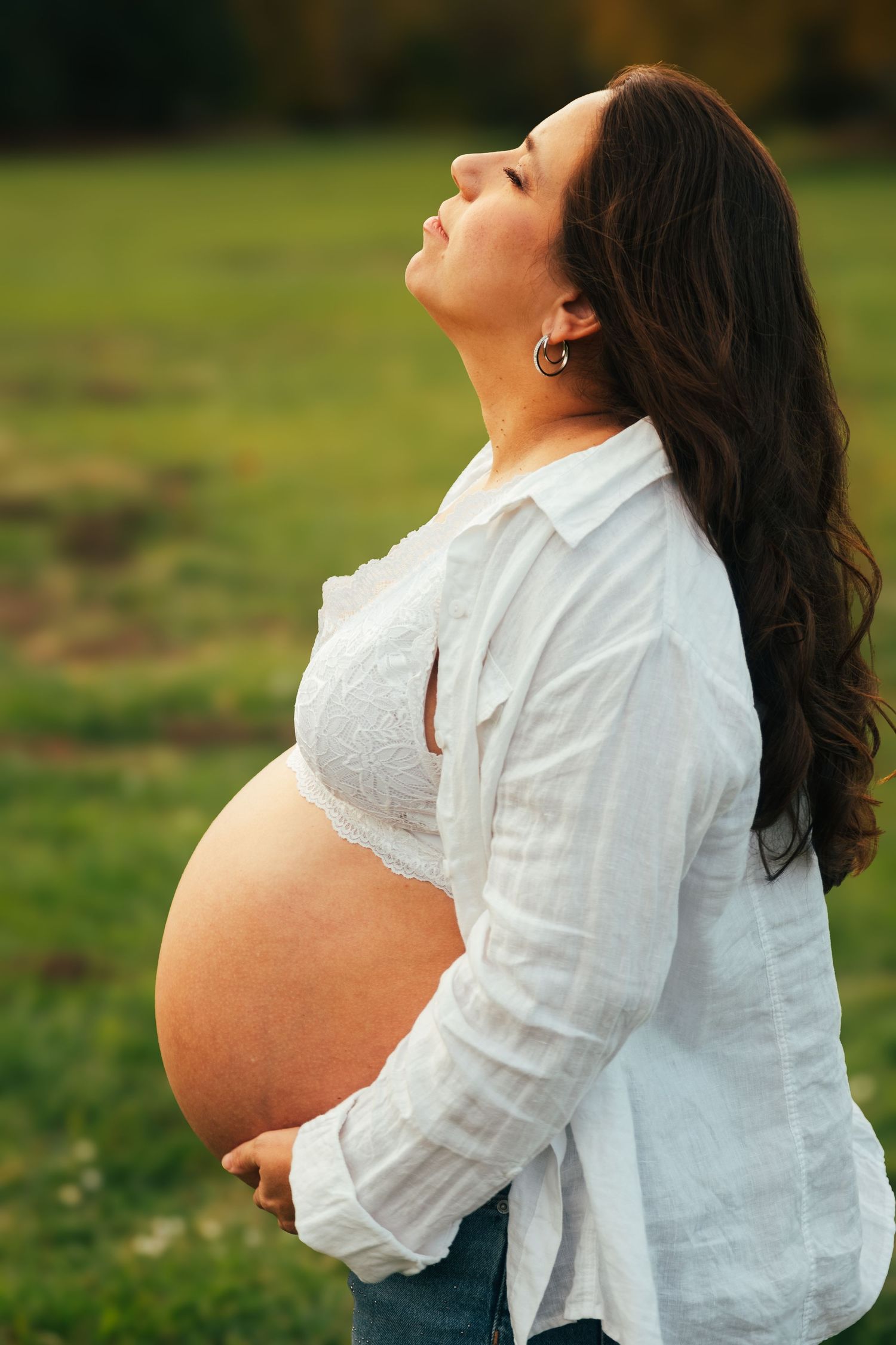 A side profile of an expectant mother in a white shirt standing in a sunlit field.