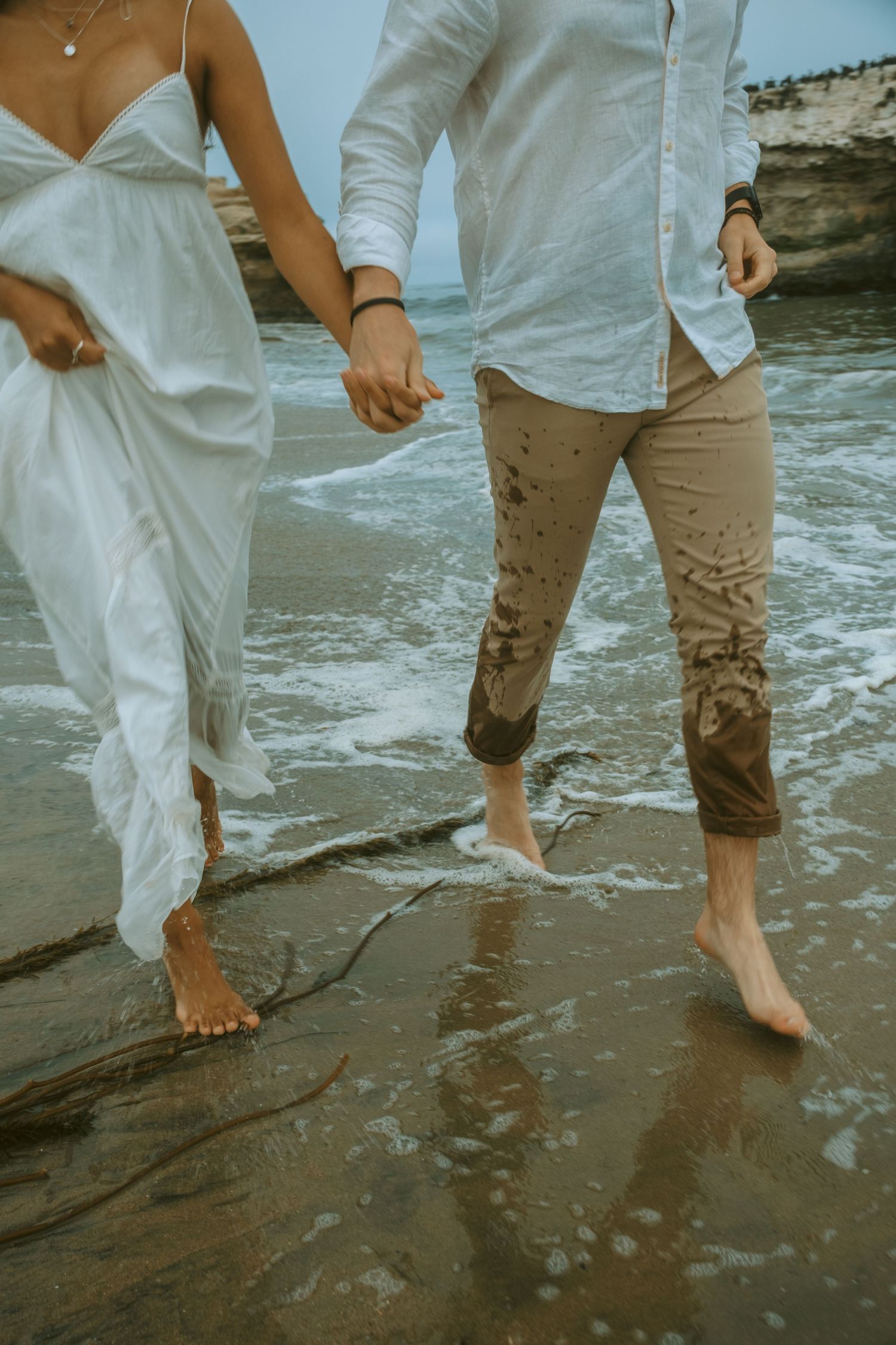 Barefoot couple walking along wet beach rocks with ocean waves splashing nearby.