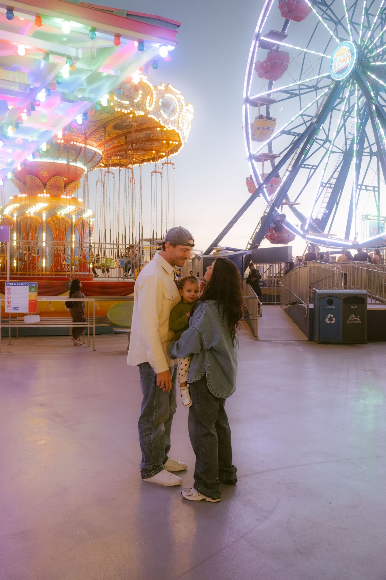 Family of three sharing a tender moment at an amusement park with a lit carousel and Ferris wheel at dusk.
