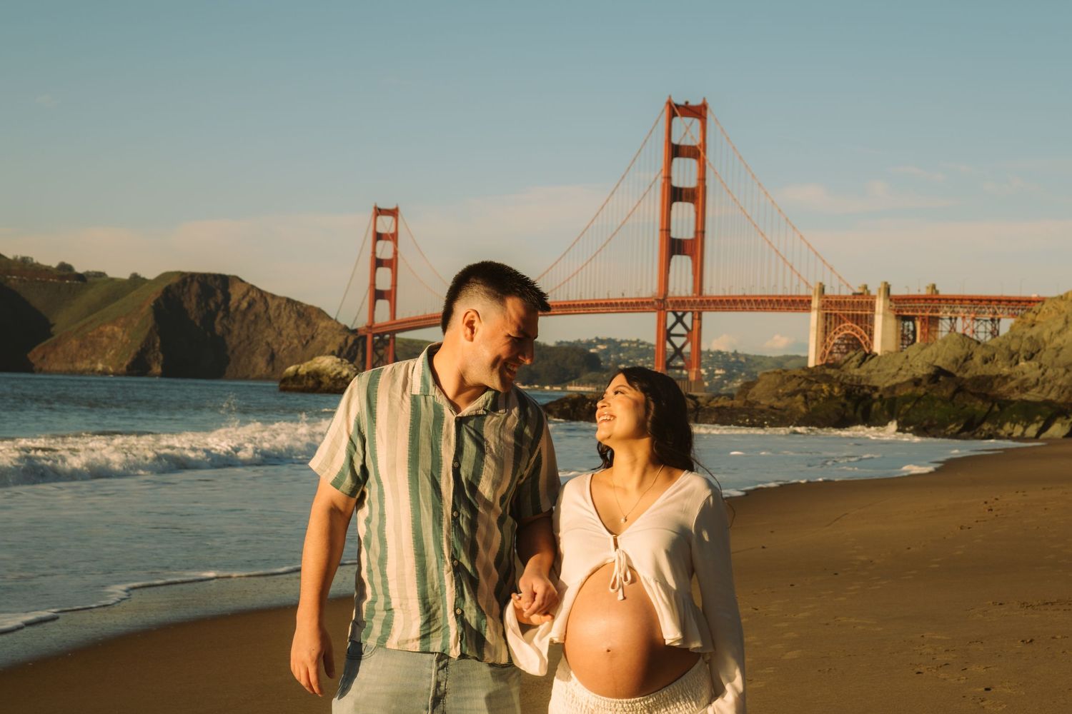 Pregnant couple holding hands on Baker Beach with Golden Gate Bridge backdrop in San Francisco.