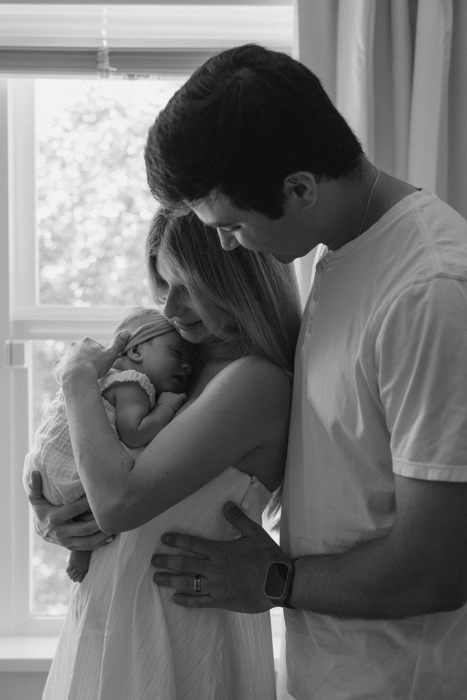 Black and white photo of new parents tenderly cradling their newborn baby by a bright window.