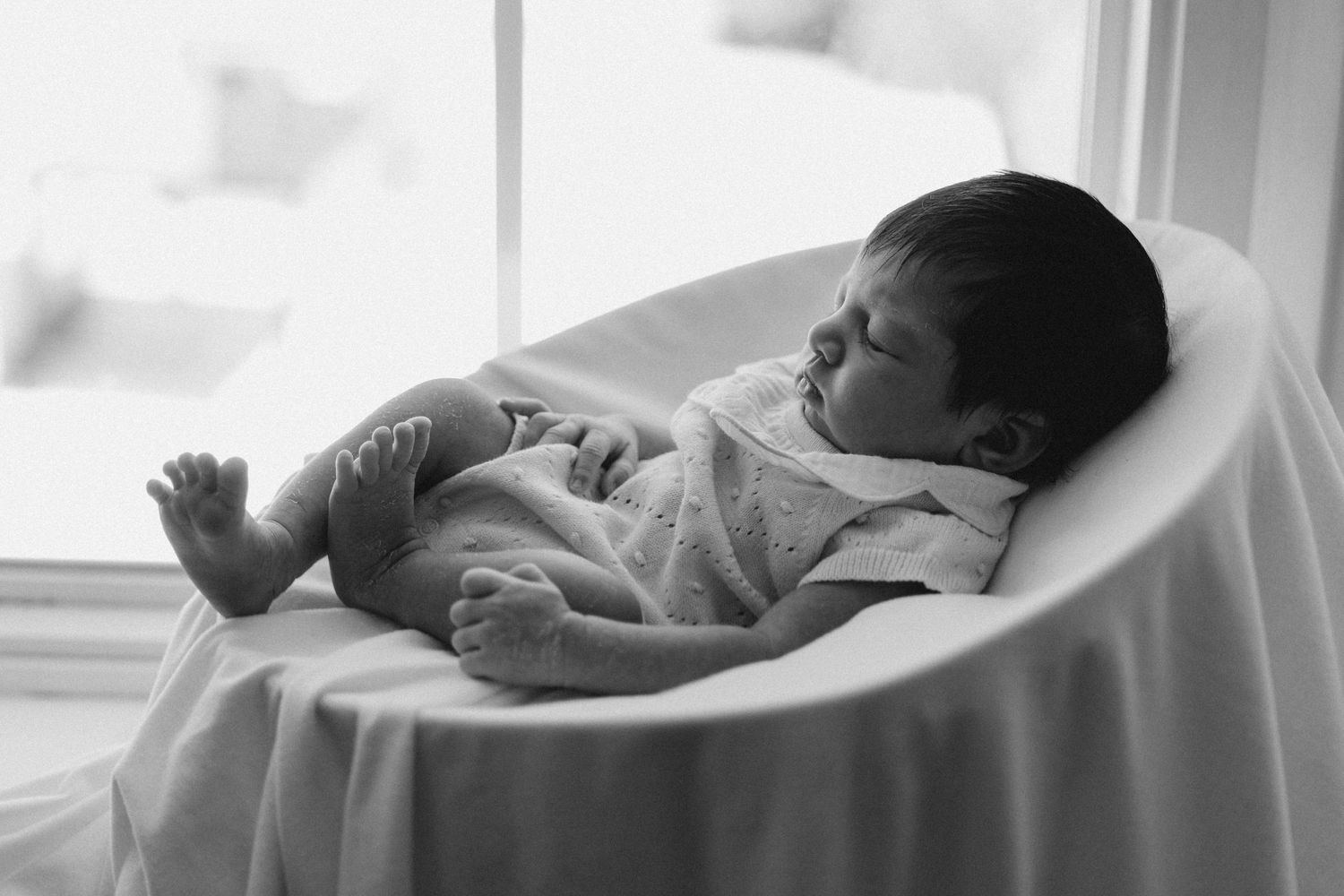 Black and white photo of a sleeping newborn baby curled up peacefully in a white bowl-shaped seat near a window.