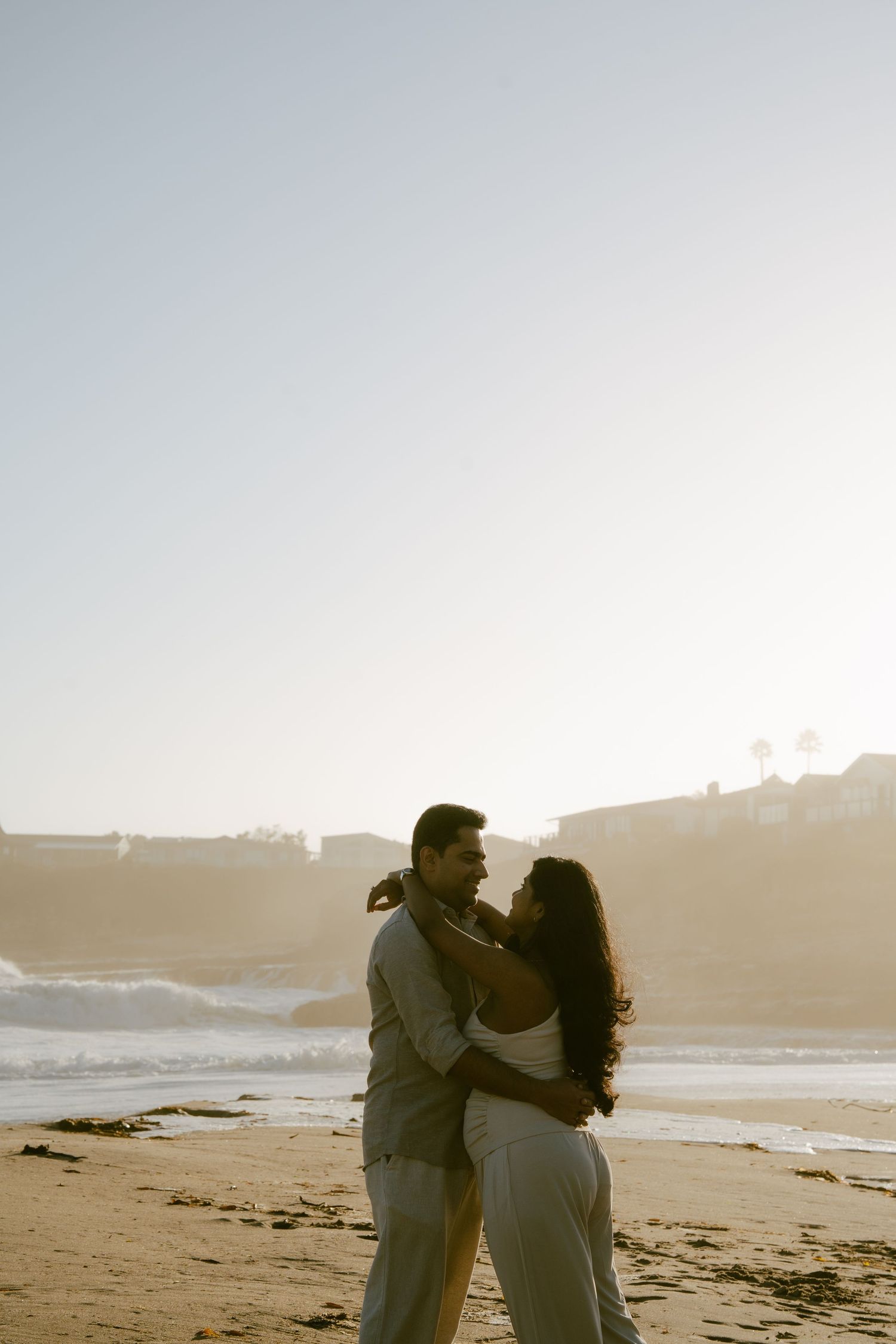 Couple embracing romantically on a misty beach at sunset with ocean waves and coastal cliffs in background.