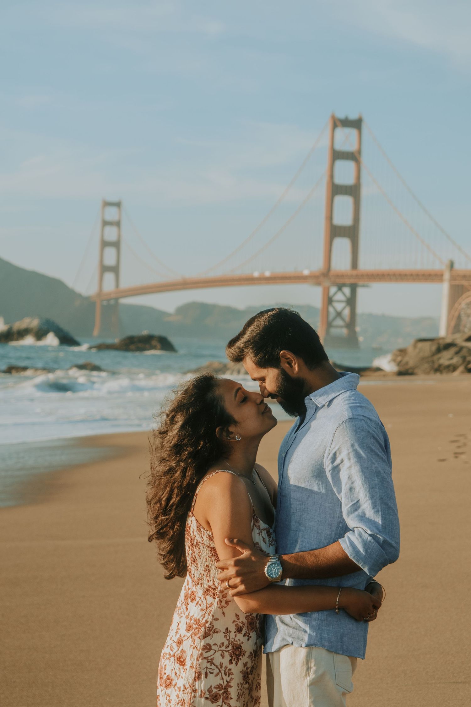 Couple embracing on Baker Beach with the iconic Golden Gate Bridge in the background on a sunny day.