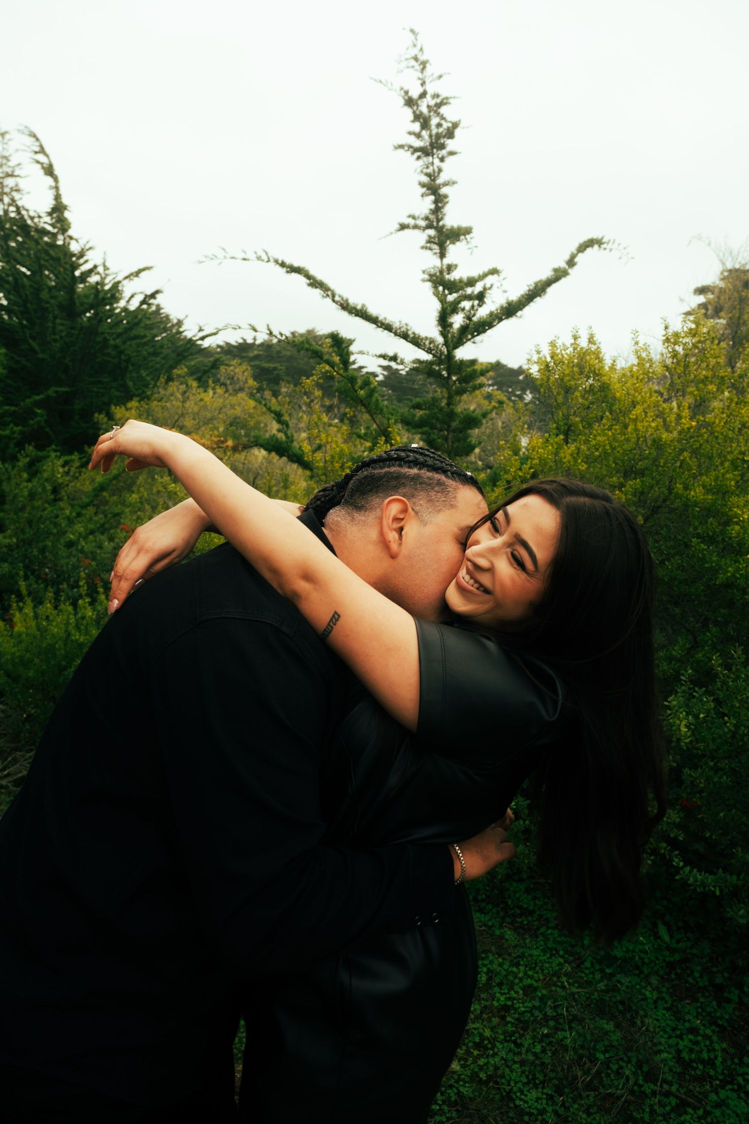Couple embracing and smiling in a lush green garden, wearing matching black outfits.