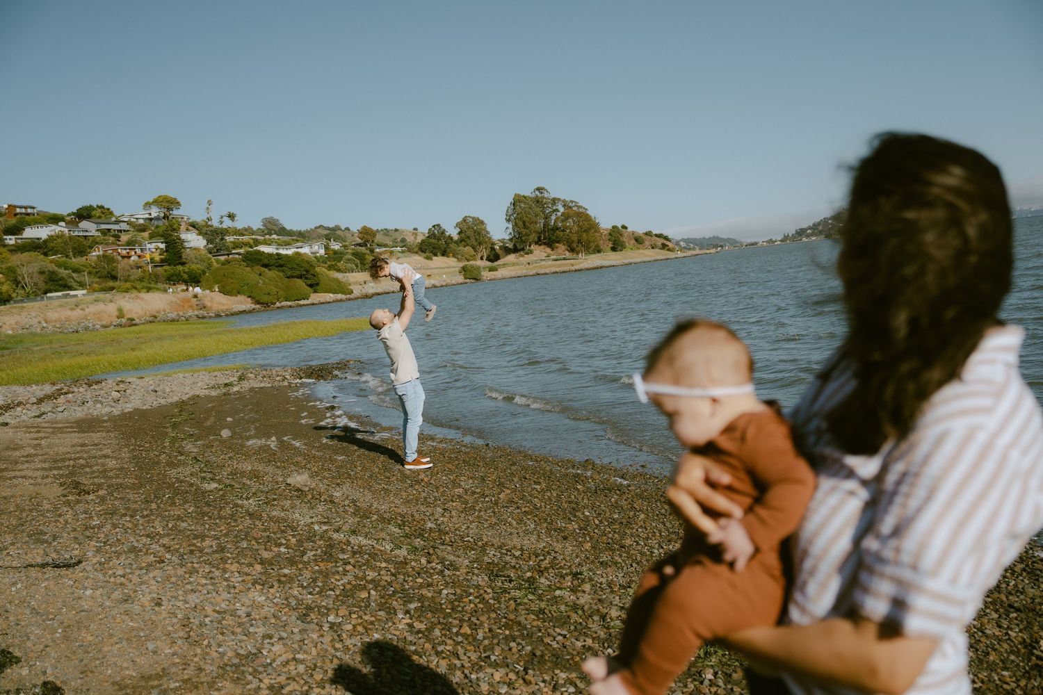Family enjoying a sunny day by the river, with a woman holding a baby and another person skipping stones.