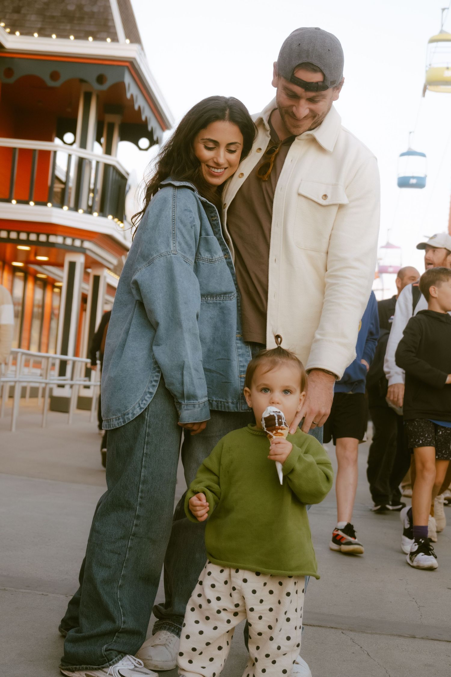 A young family with a toddler eating ice cream strolls at an amusement park, parents dressed casually in denim and white.
