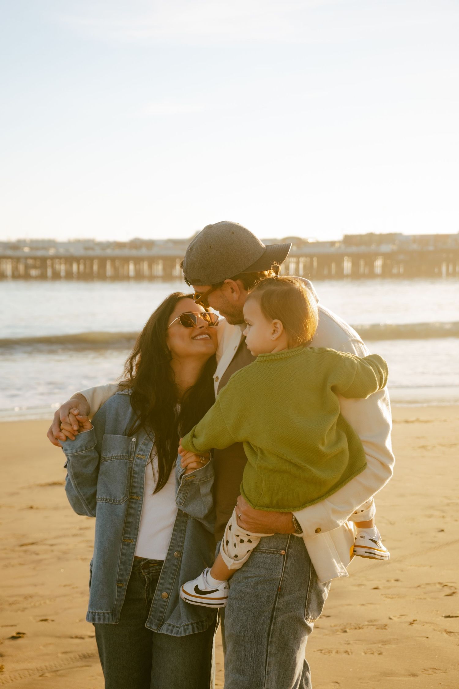 Young family sharing a tender moment on a sunny beach, parents kissing their toddler near a pier.