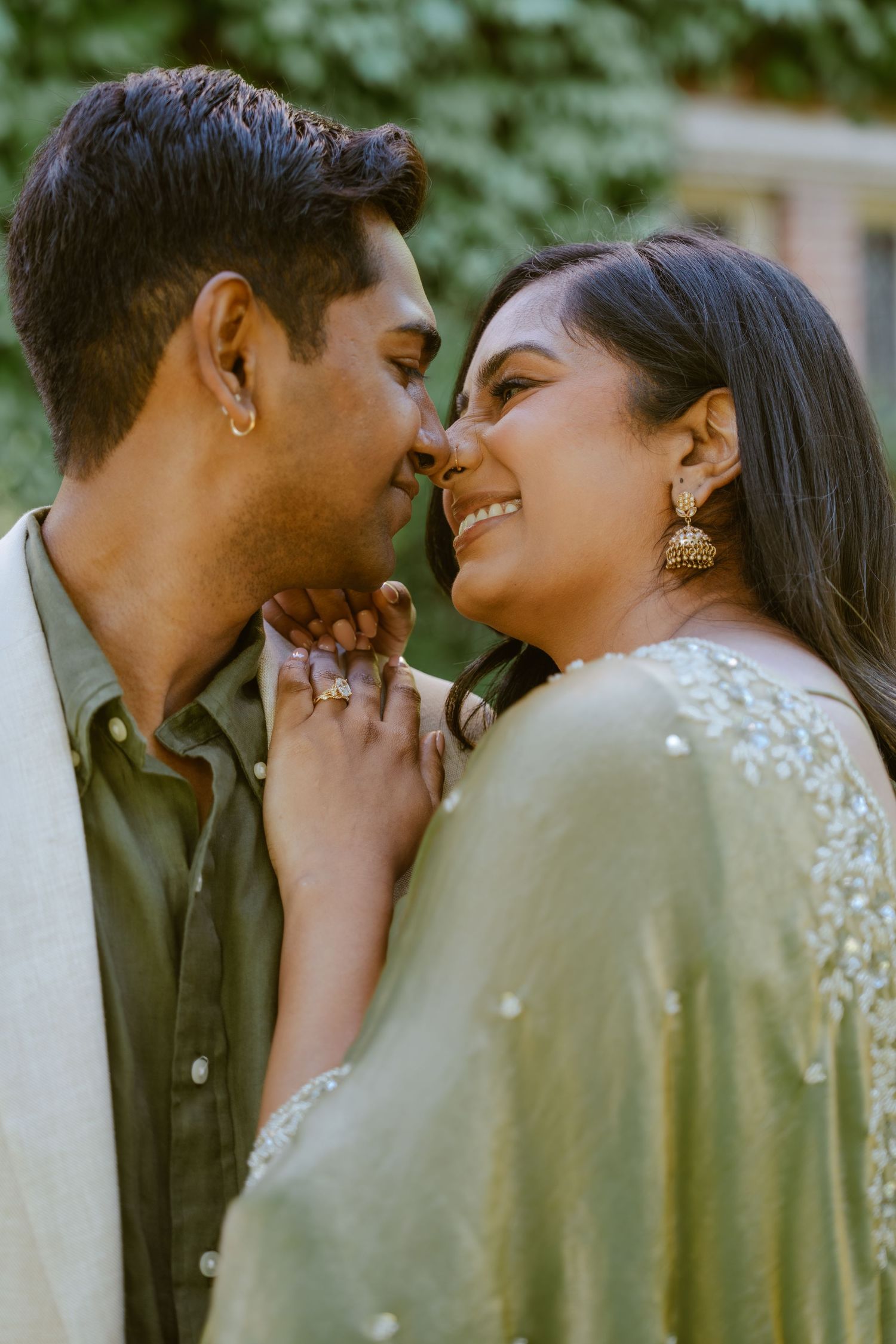A couple in traditional South Asian attire share a romantic moment, smiling at each other outdoors.