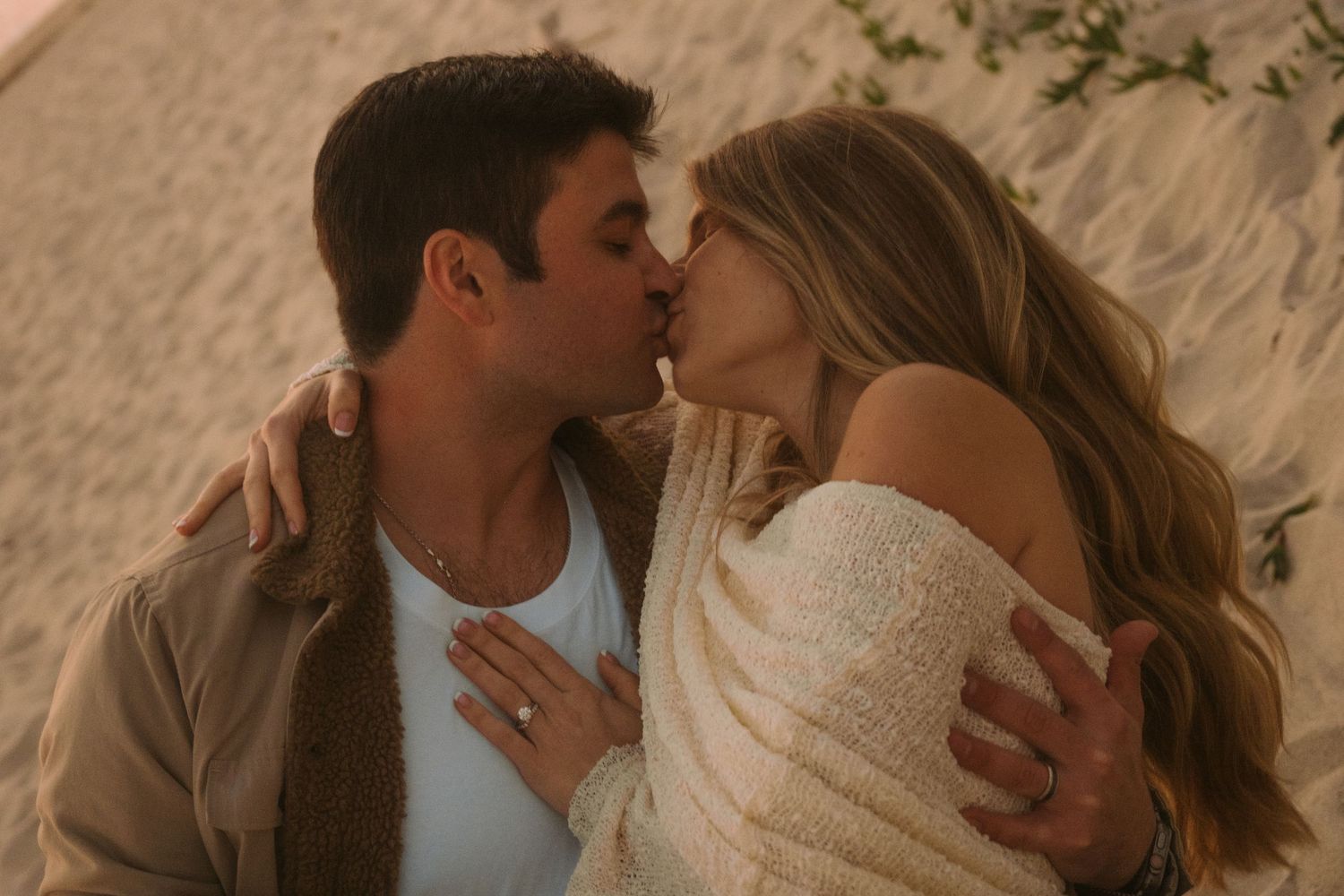 Couple sharing a romantic kiss on a sandy beach, woman wearing an engagement ring and off-shoulder cream sweater.