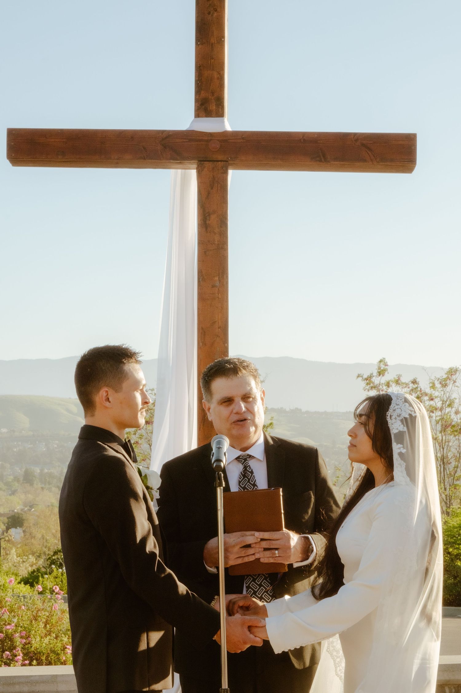 Couple exchanging vows at outdoor wedding ceremony before large wooden cross with scenic mountain views.