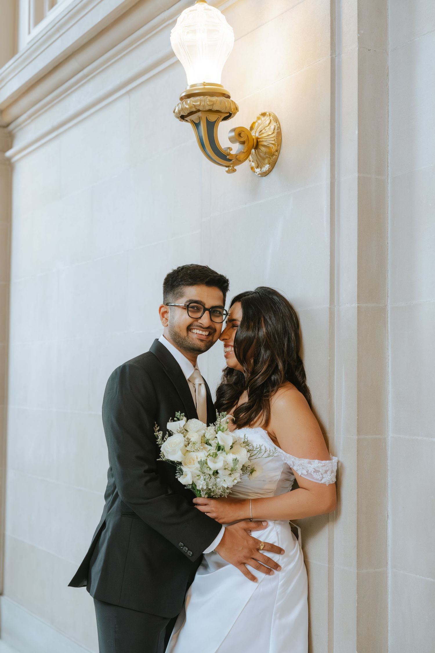 Smiling groom in dark suit embraces bride in white off-shoulder gown holding bouquet, posing by elegant wall sconce.