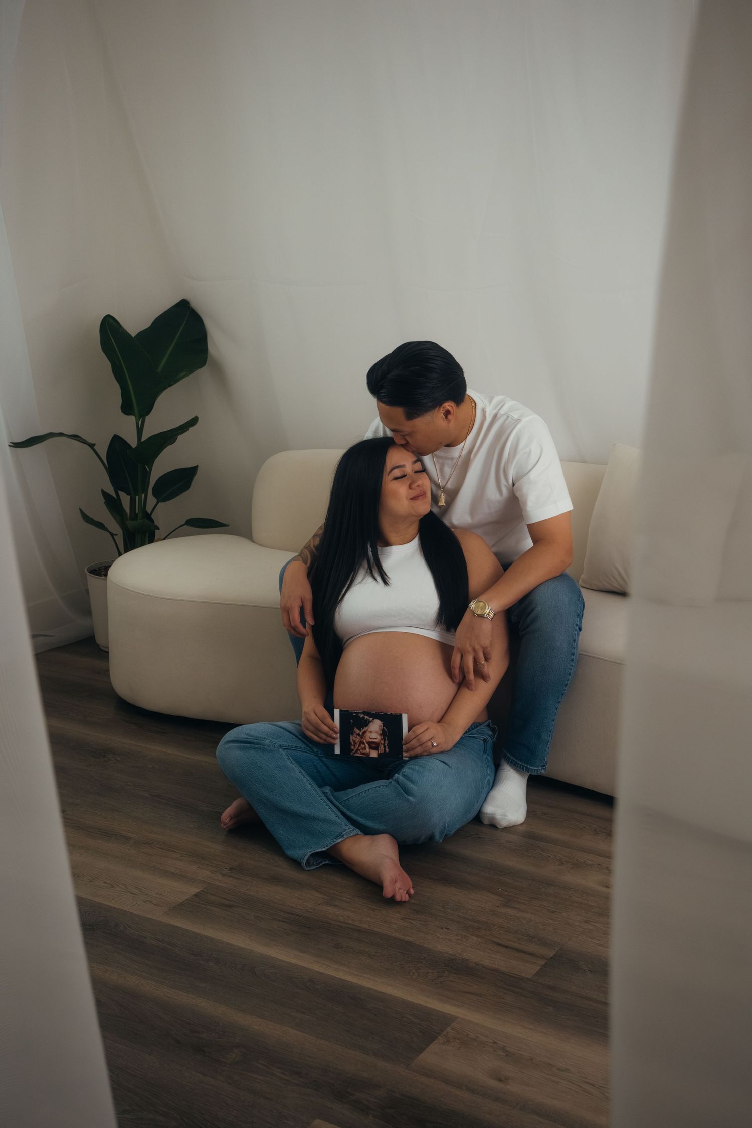 Couple shares tender moment on white couch during maternity photoshoot with indoor plant in background.