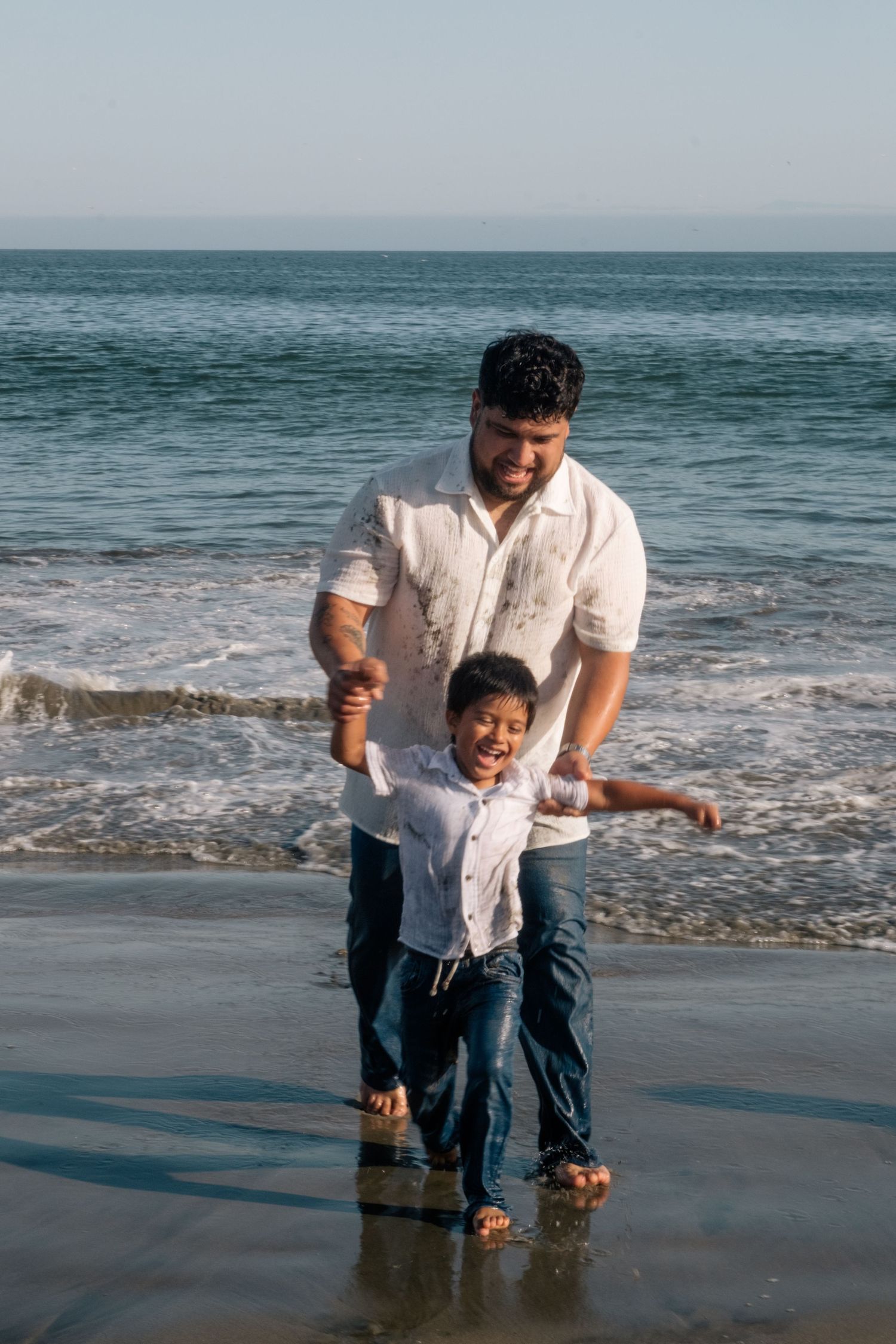 Father and young son laughing and playing together at the beach shore, both wearing white shirts and jeans.