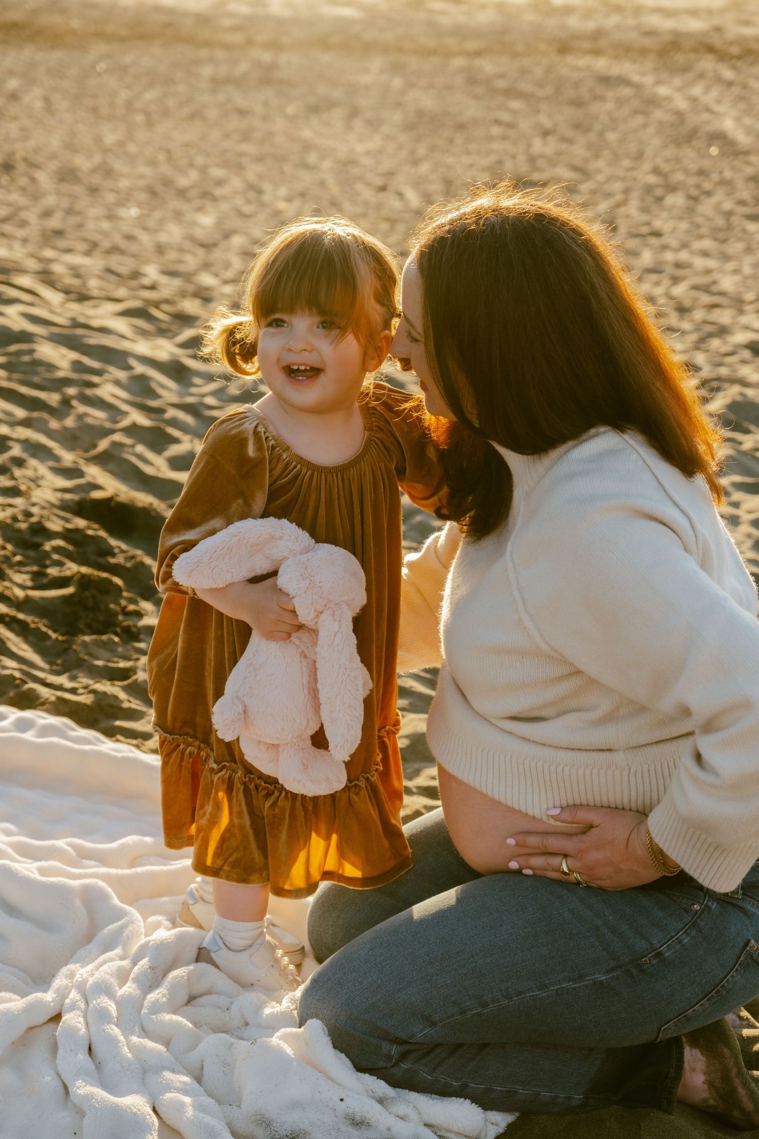 Pregnant woman kneeling on beach blanket with toddler girl holding pink bunny at golden hour sunset.