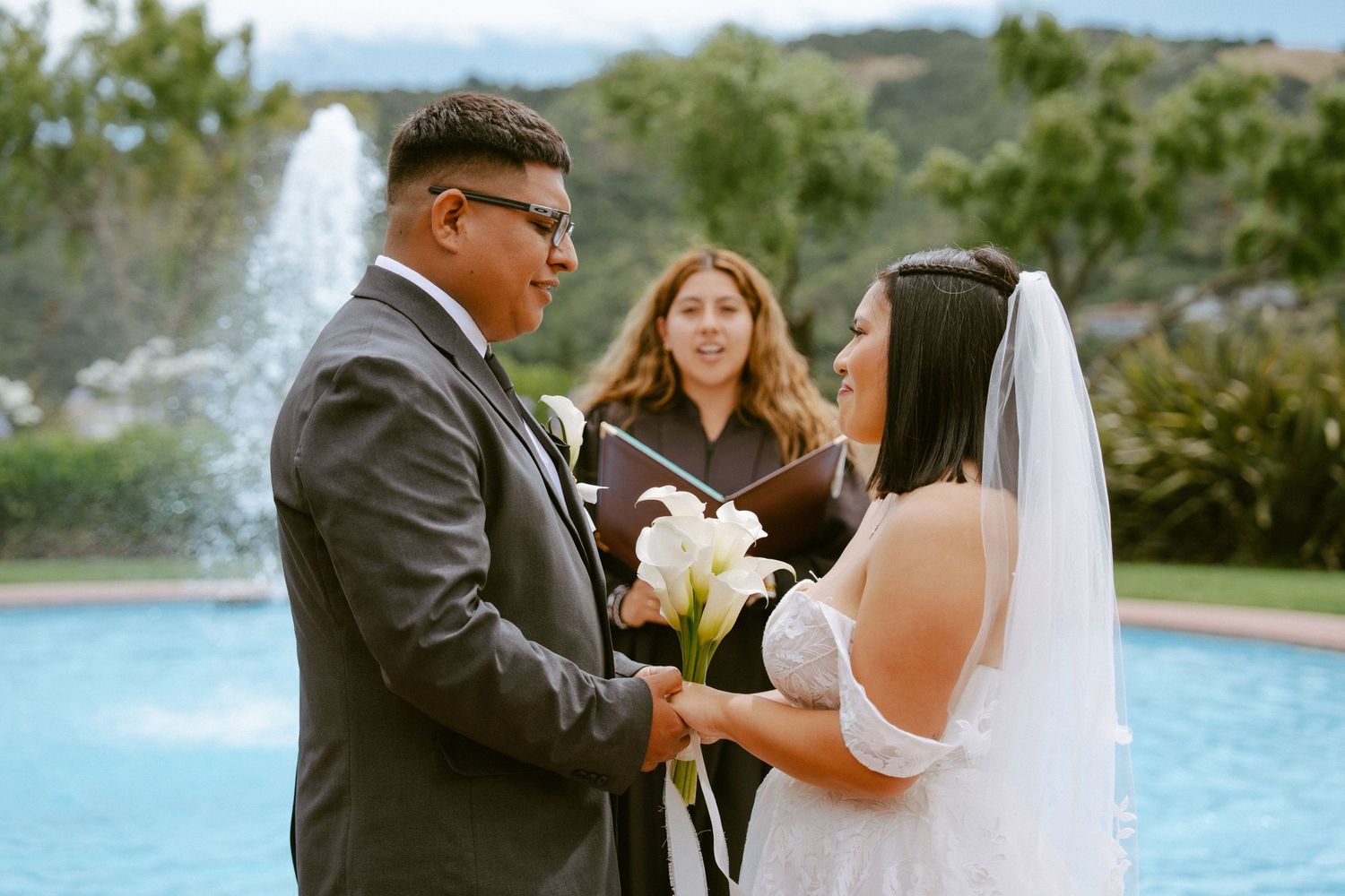Bride and groom exchange vows outdoors by a fountain with an officiant, bride holding white calla lilies.