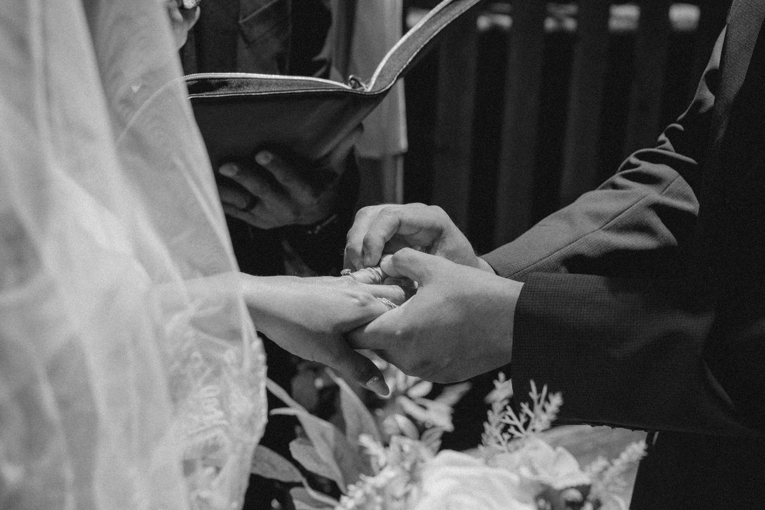 Black and white wedding ceremony photo of groom placing ring on bride's finger with officiant holding book.